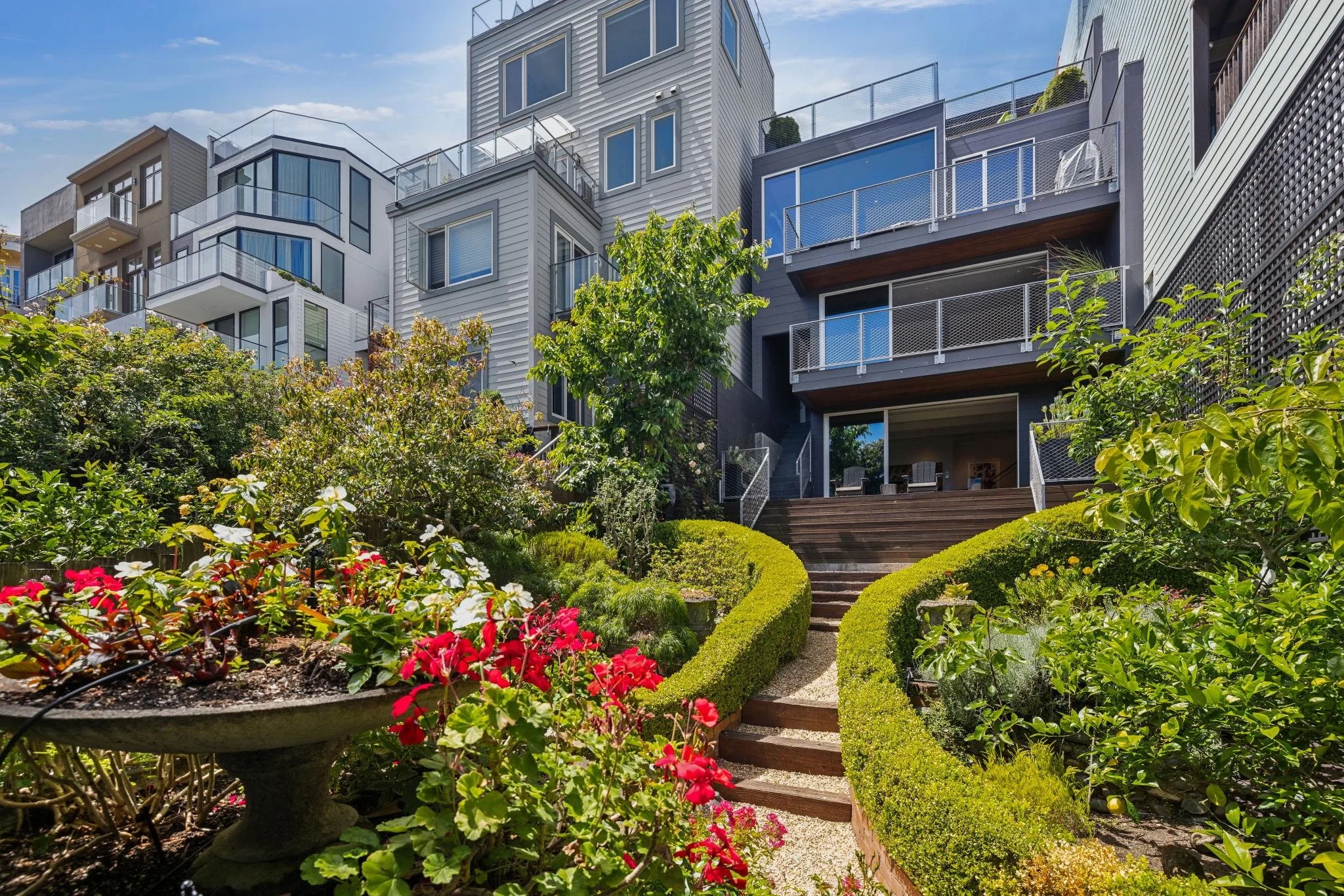 A landscaped garden with colorful flowers and neatly trimmed bushes, leading to a modern multi-story apartment complex with glass balconies.