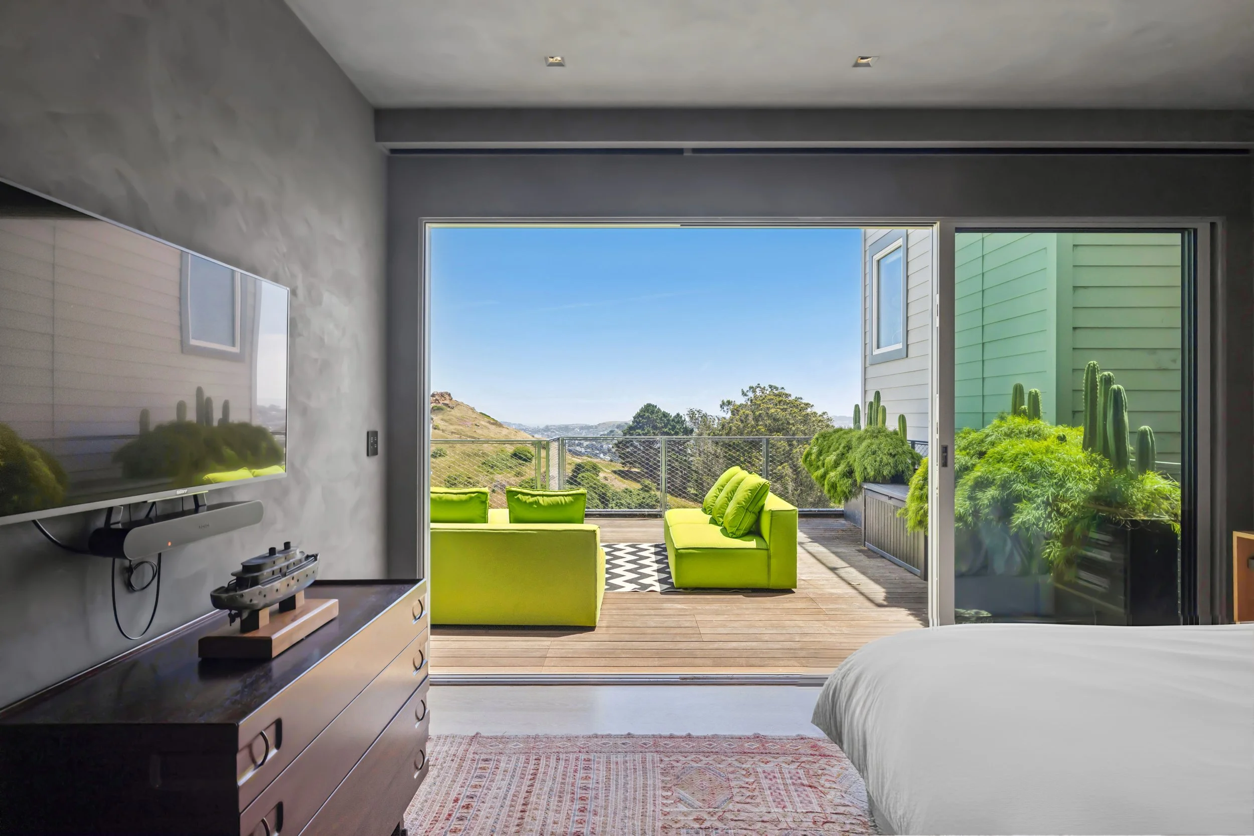 A bedroom with an open sliding glass door leading to a balcony with green outdoor furniture. The room has a dresser, a wall-mounted TV, and a bed.
