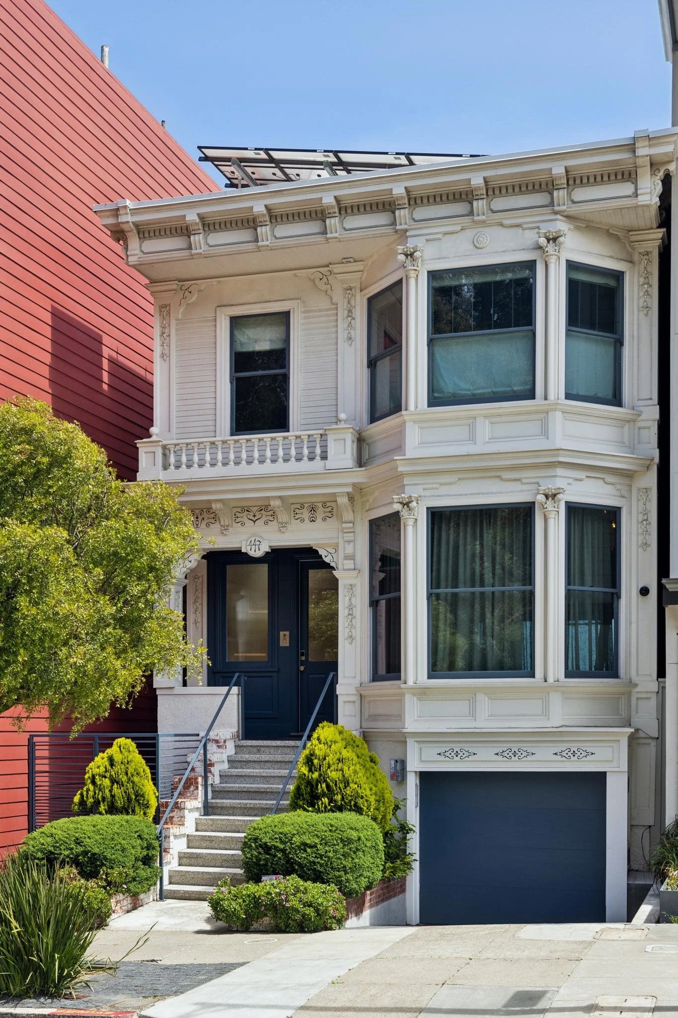 A Victorian-style house with intricate white woodwork, several large bay windows, and a dark blue front door. The house has stairs leading up to the entrance, surrounded by well-maintained bushes and green plants. There is a large green tree to the l