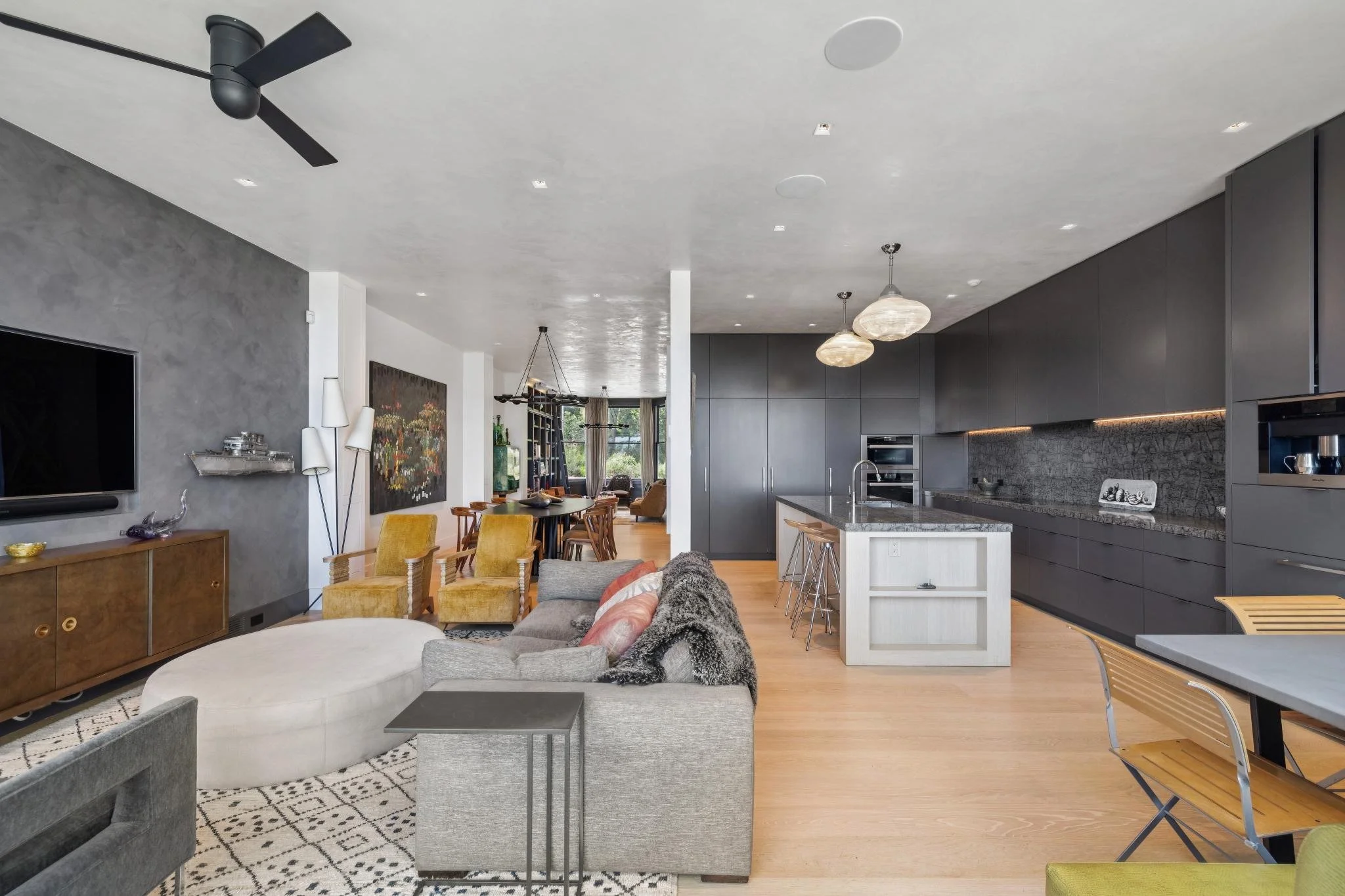 Open-concept living room and kitchen with modern decor, gray cabinets, pendant lighting, and a dining area with wooden chairs.