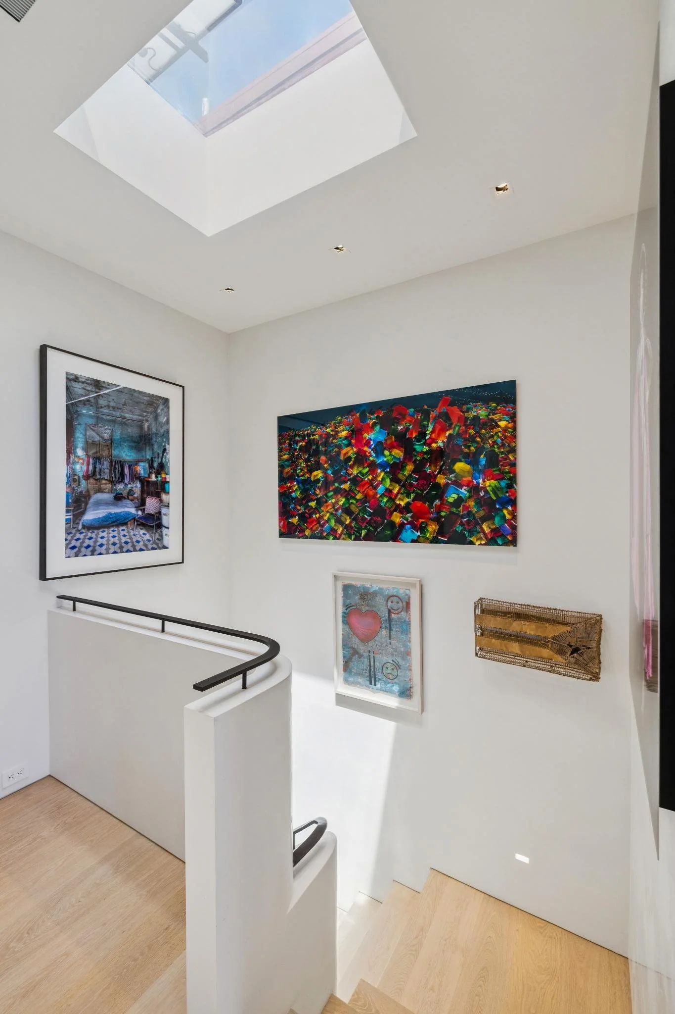 Interior of an art gallery with paintings on white walls, a staircase with a black handrail, and a skylight on the ceiling.