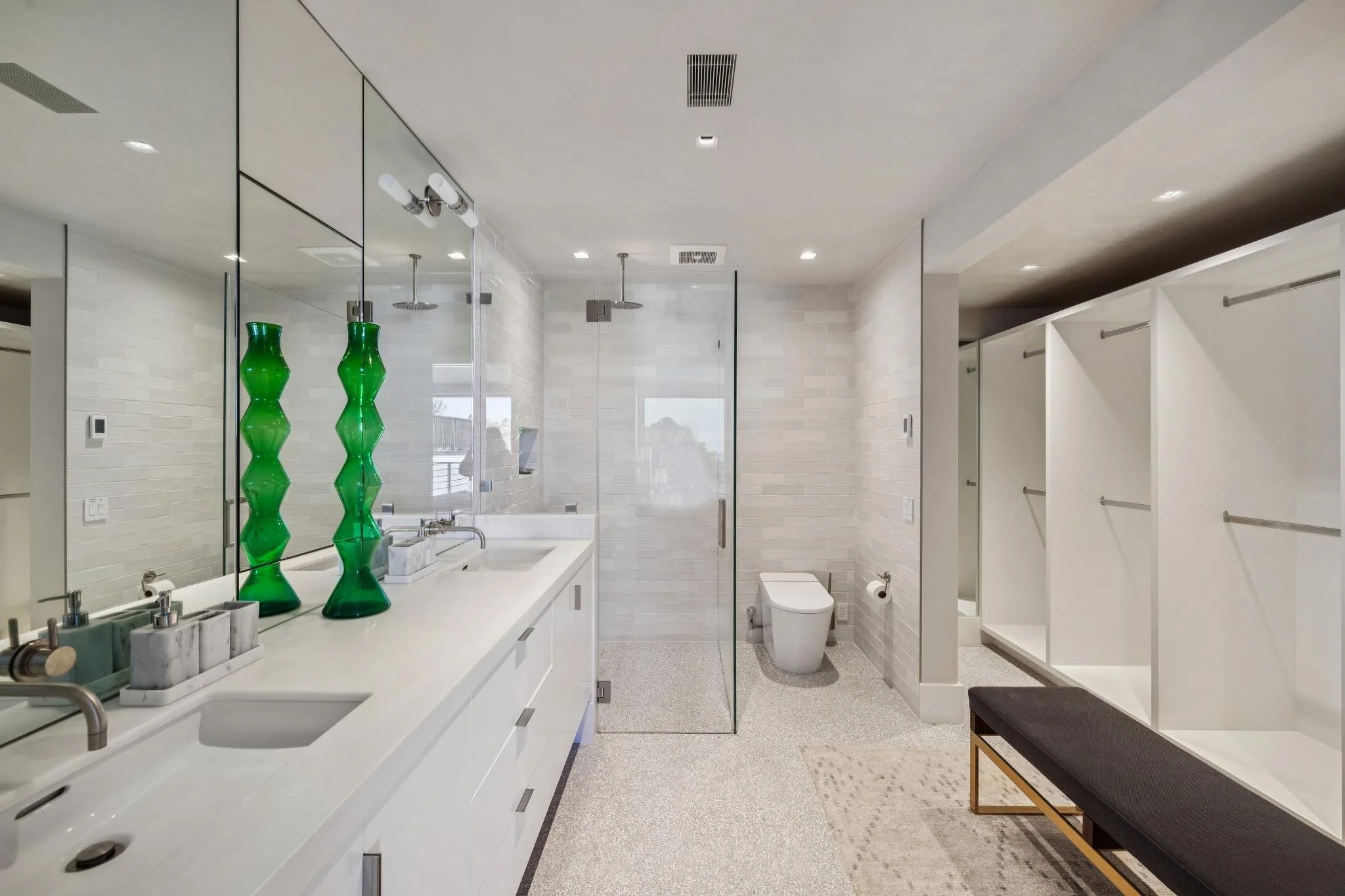 Modern minimalist bathroom with white cabinets, green vases, glass shower enclosure, towel hooks, bench, and a toilet.