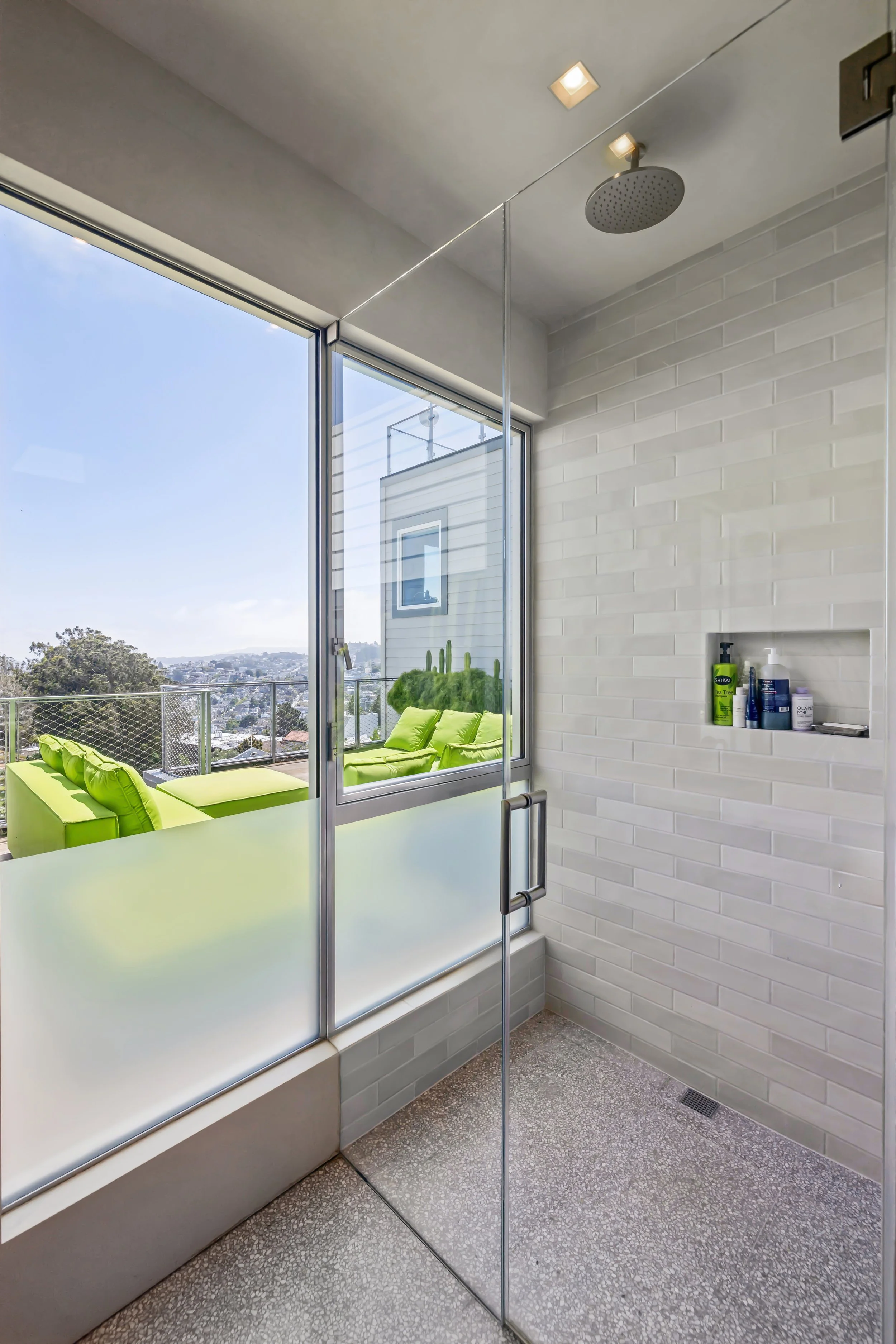 Balcony with lime green outdoor furniture, large windows, view of cityscape, white brick wall, personal care products on a ledge, ceiling light, rain shower head.