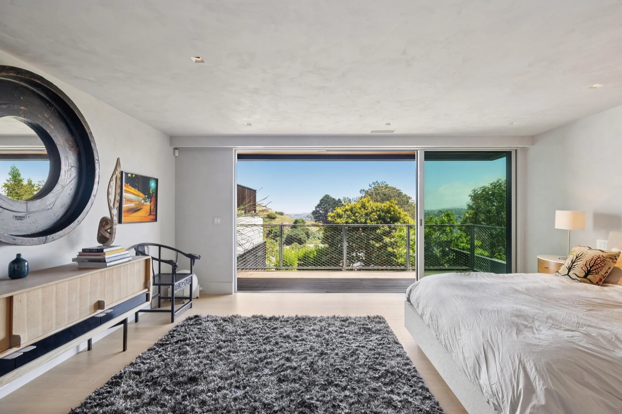 Interior of a modern bedroom with a sliding glass door leading to a balcony, a white bed with decorative pillows, a black and white shaggy rug, a light wood sideboard with books and decorative items, a flat-screen TV on the wall, and a lamp, with tre