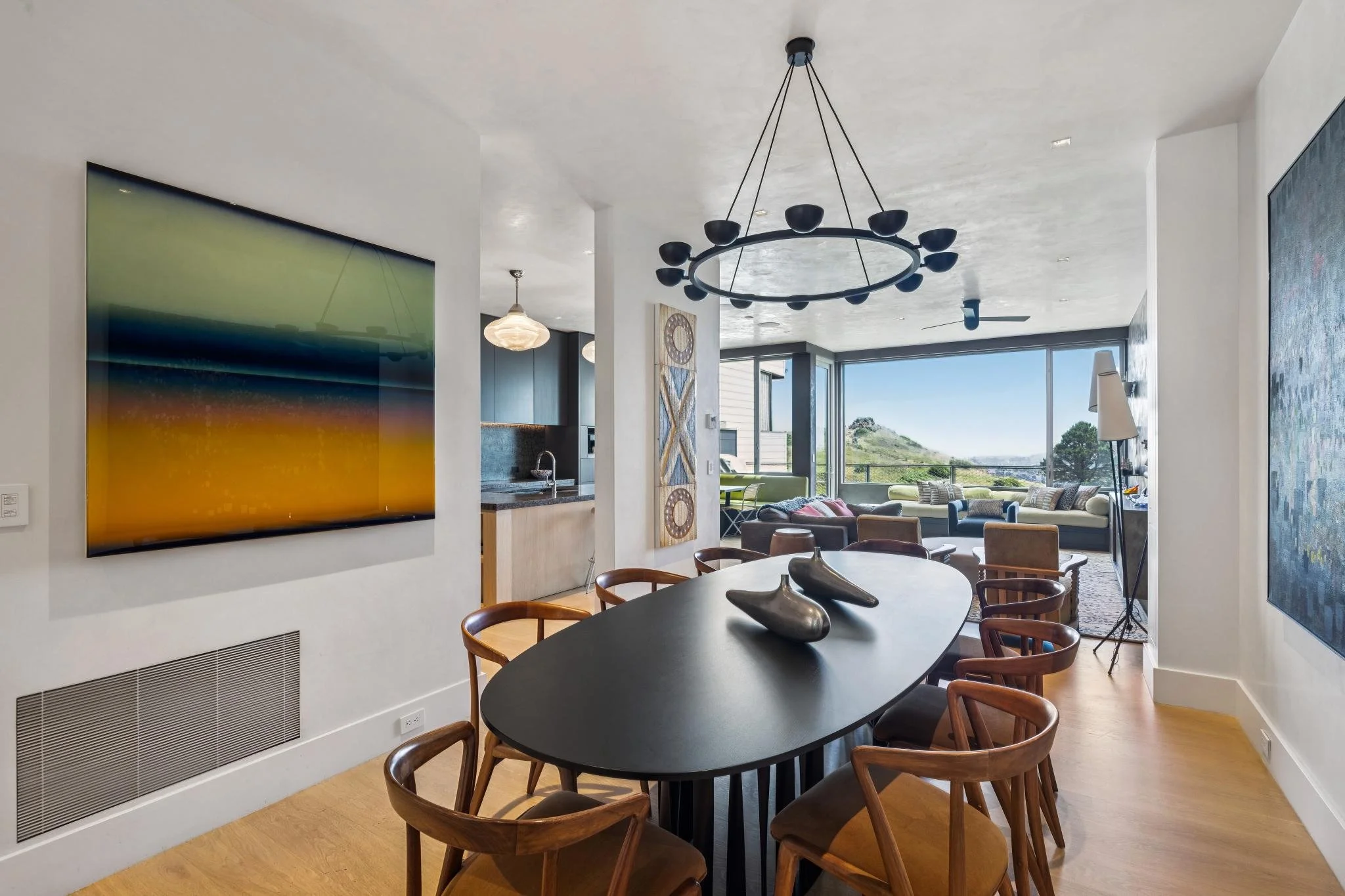 Modern open-concept living and dining area with a black oval dining table, wooden chairs, contemporary artwork, large window with a view of greenery, ceiling fan, and a circular chandelier.