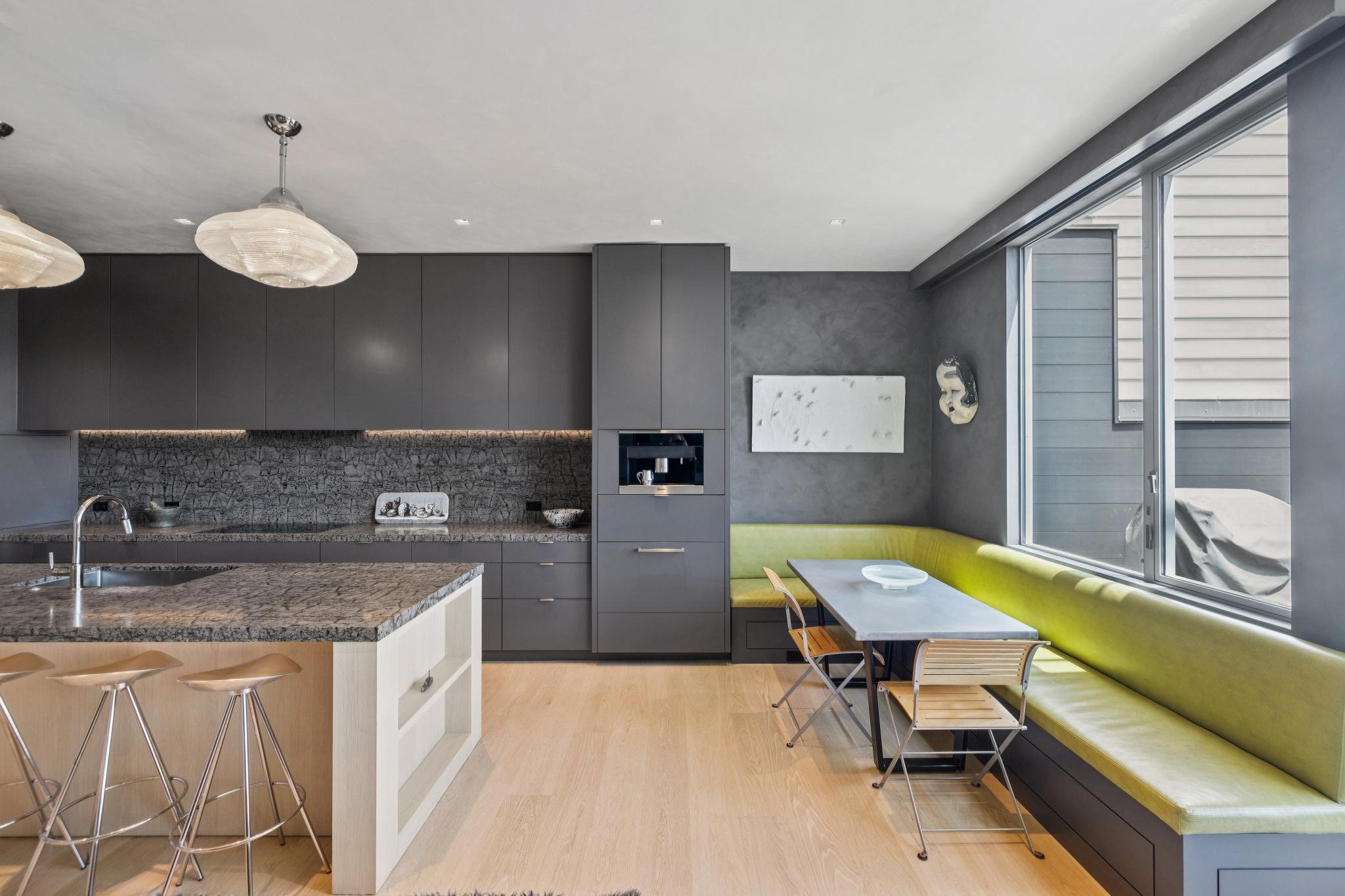 Modern kitchen and dining area with gray cabinetry, a granite countertop, a built-in coffee machine, a marble backsplash, a white kitchen island with barstools, and a nook with a bright green cushioned seating and a window.