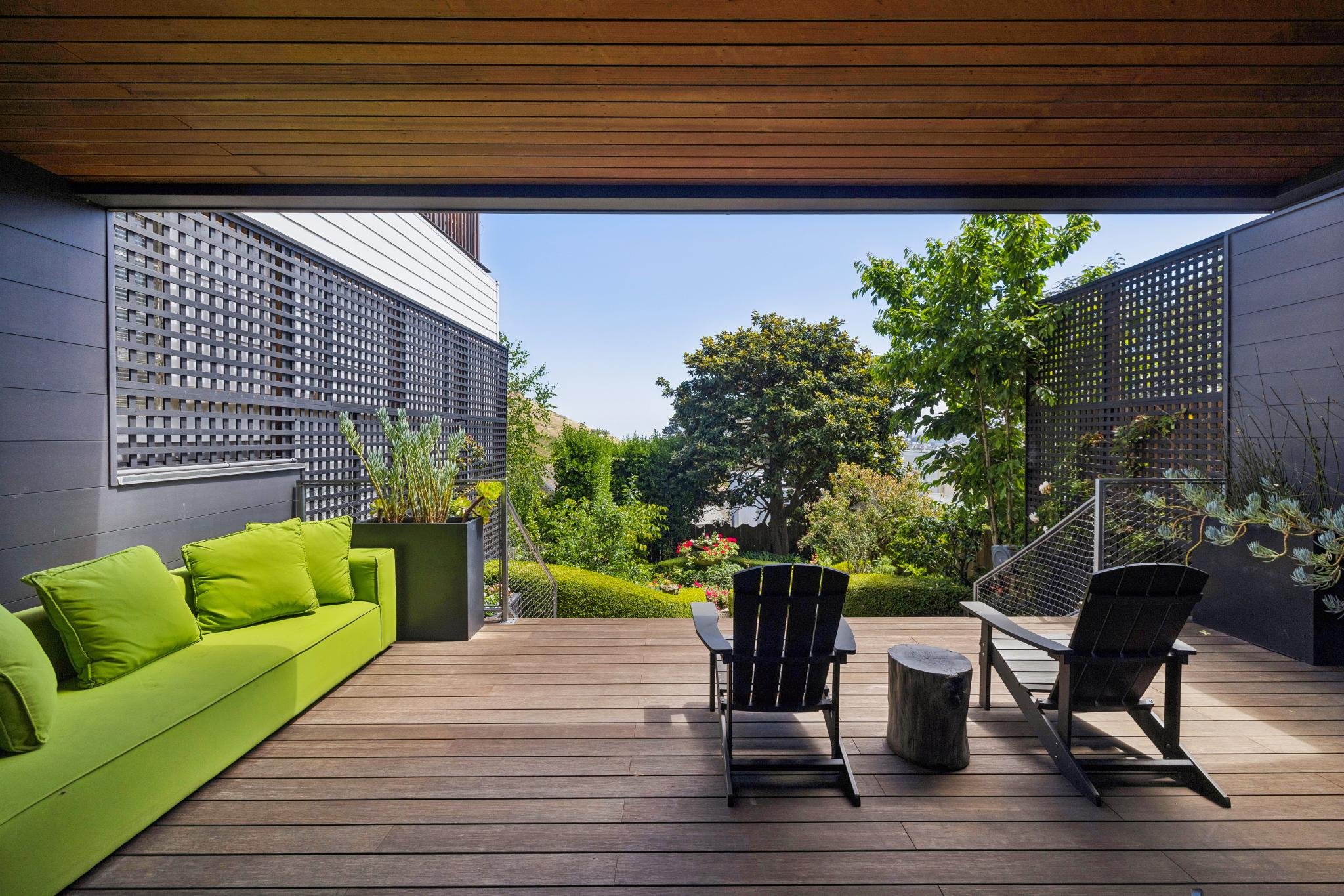 View from a balcony or patio with a lime green sofa, black chairs, and a wooden deck. In the background, there are trees and lush greenery under a clear blue sky.