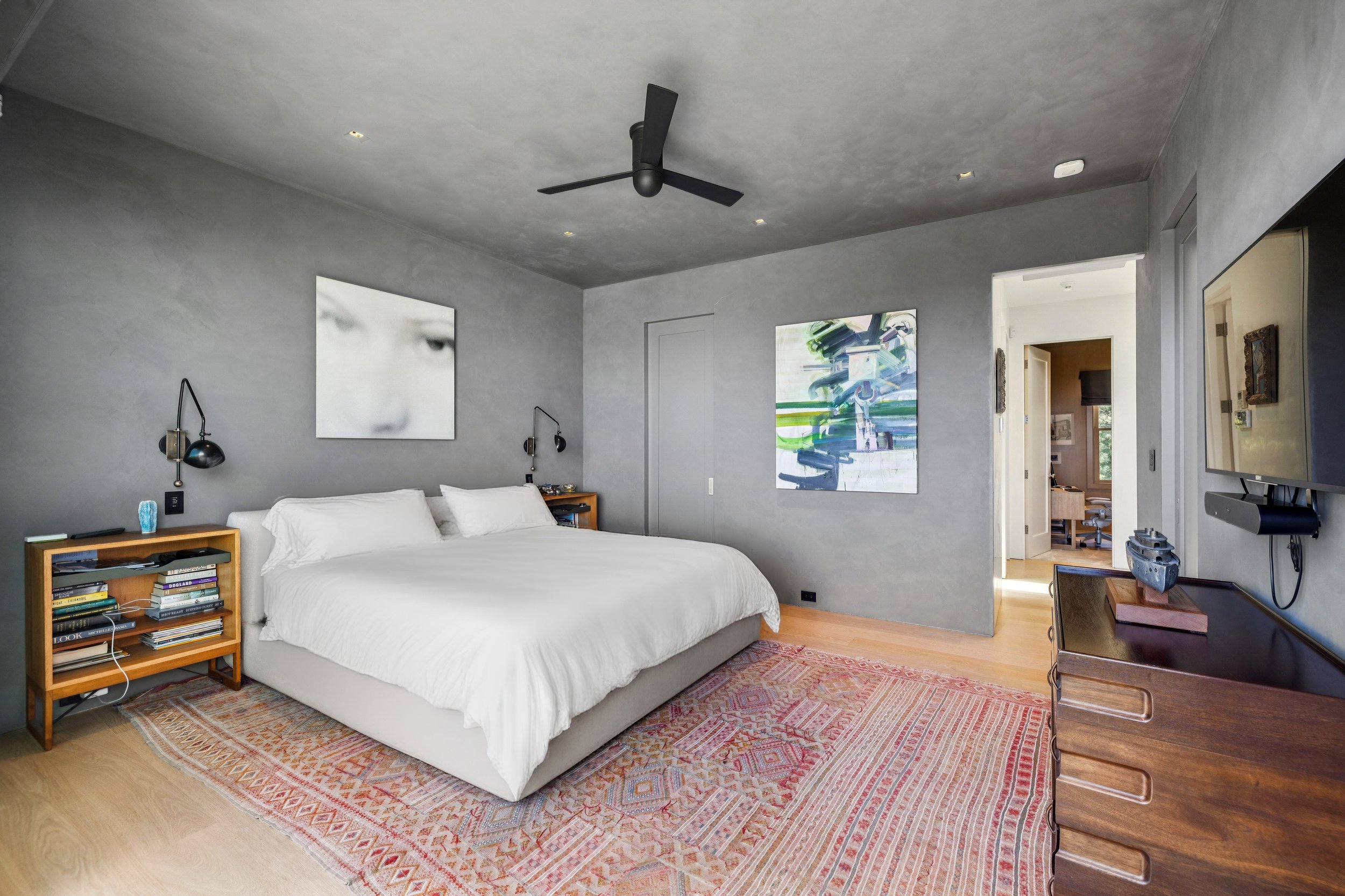 A modern bedroom with a white bed, wooden nightstands, abstract art on the walls, a ceiling fan, a television, and a patterned rug on light-colored wooden floors.
