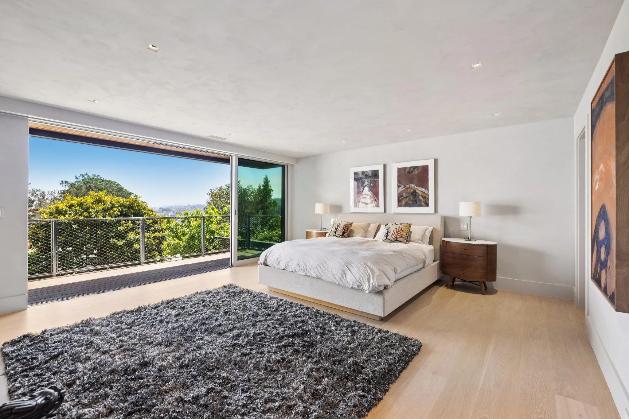 A modern bedroom with a large sliding glass door opening to a balcony surrounded by greenery, featuring a white bed with decorative pillows, side tables with lamps, and abstract artwork on the walls.