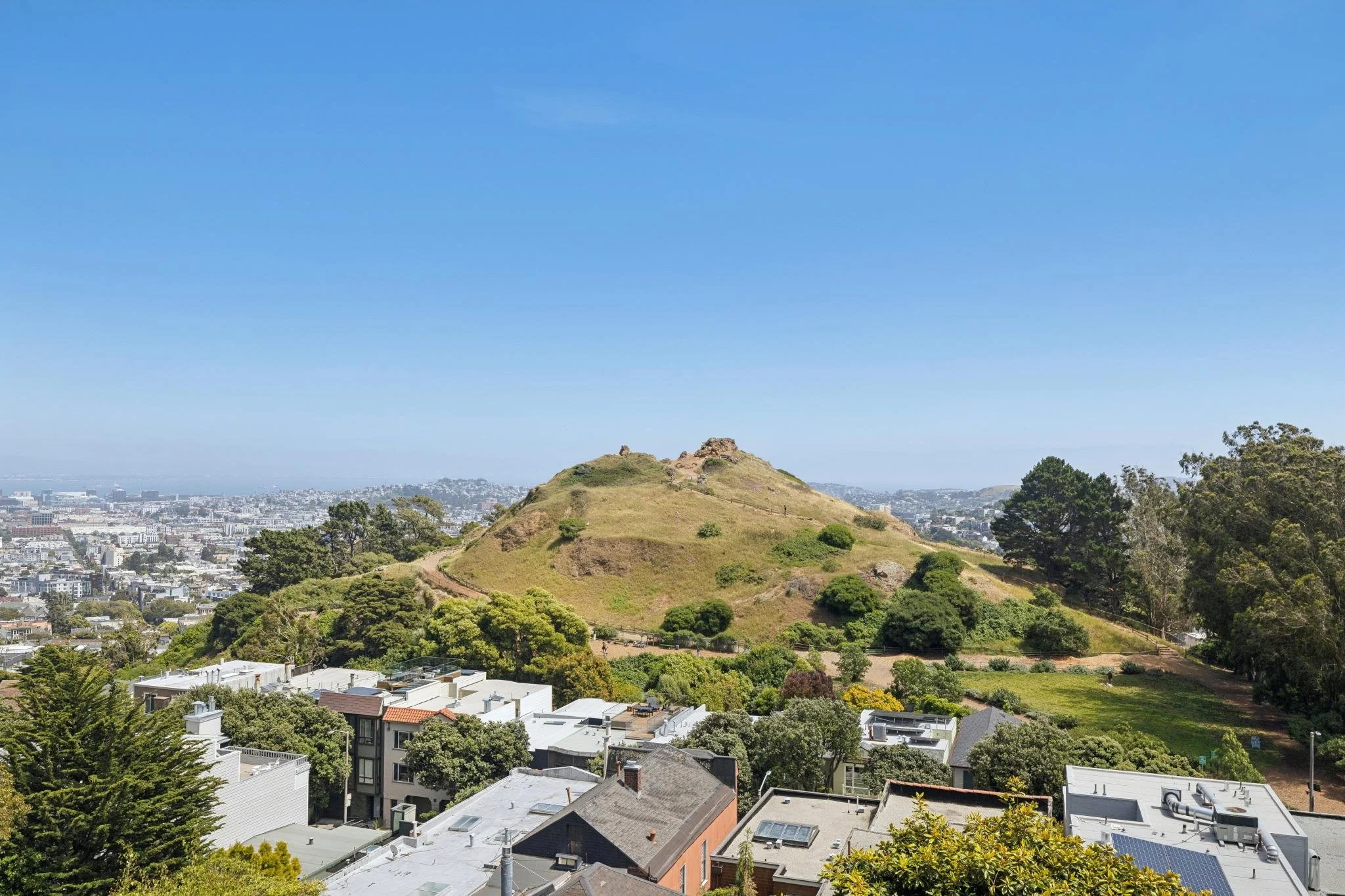 A scenic view of a city with a prominent green hill or small mountain in the center, surrounded by trees and residential buildings, under a clear blue sky.