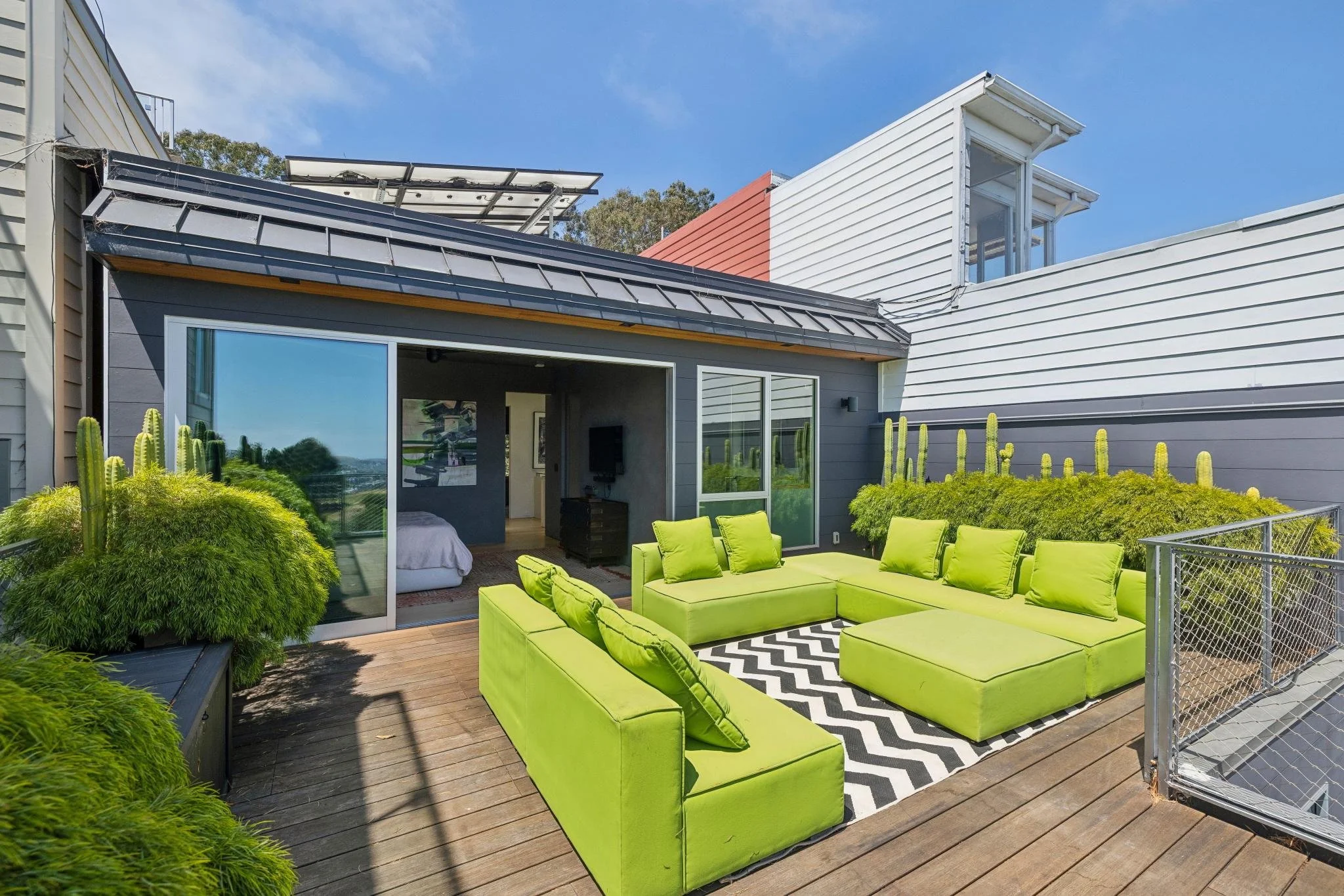 A modern house with a rooftop patio featuring bright green outdoor furniture, large windows, and plants, under a blue sky.
