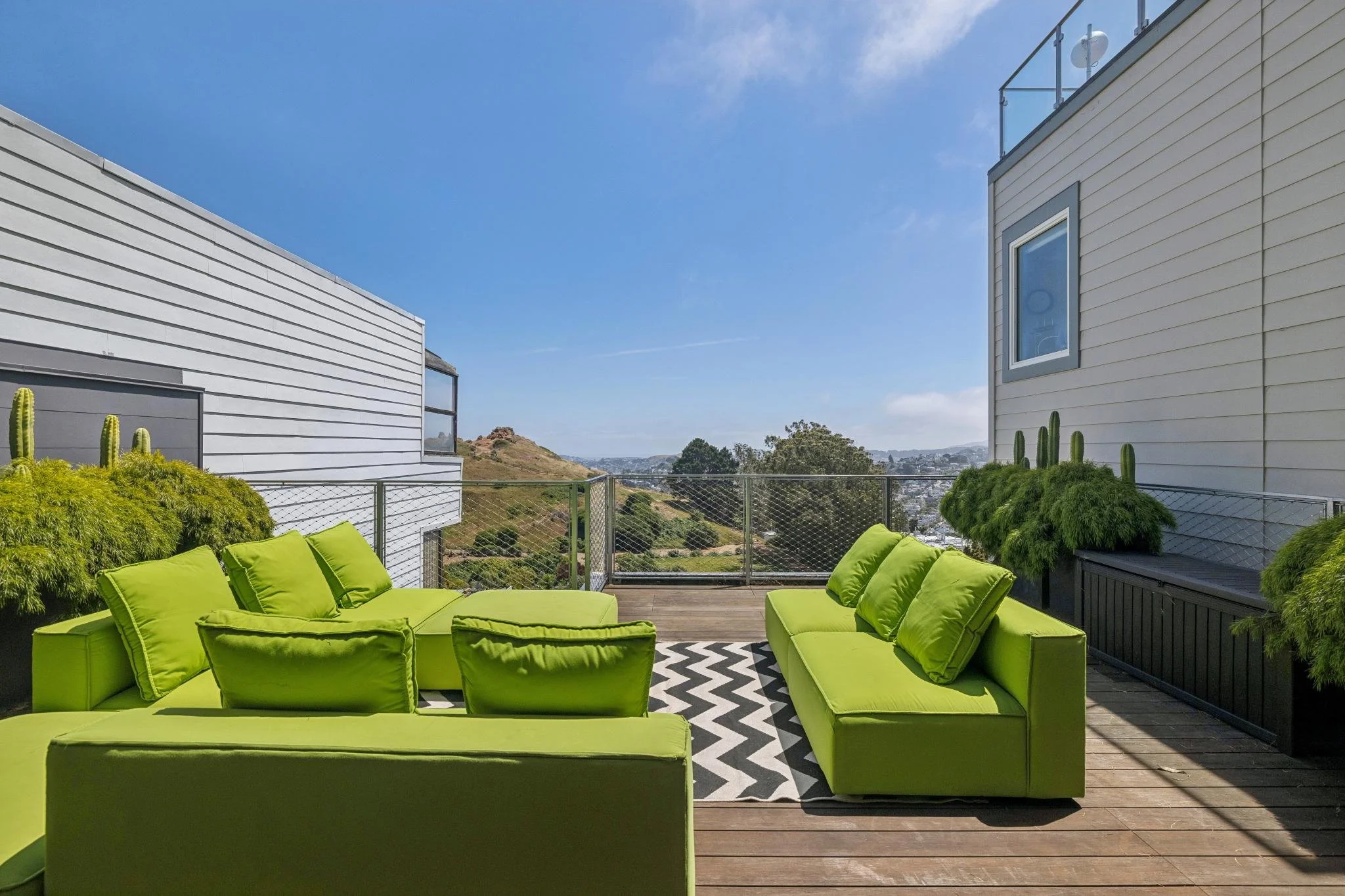A rooftop patio with bright green outdoor furniture, black and white chevron rug, surrounded by plants, with a view of rolling hills and cityscape in the distance under a partly cloudy sky.