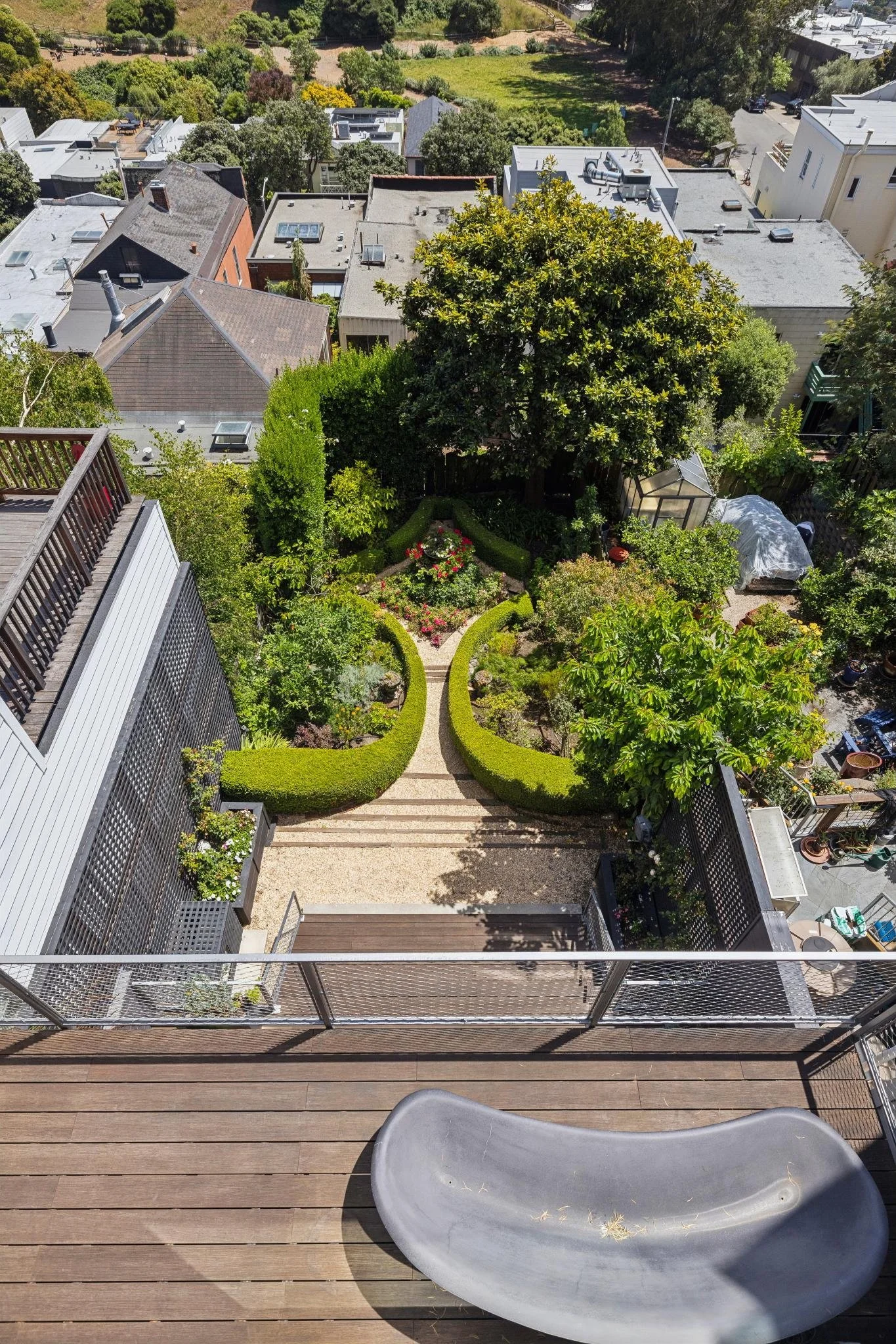Top-down view of a balcony with a gray plastic bathtub in the lower right corner, wooden floorboards, a railing with potted plants, overlooking a garden below with a curved pathway, lush trees, and surrounding houses with flat roofs and chimneys.