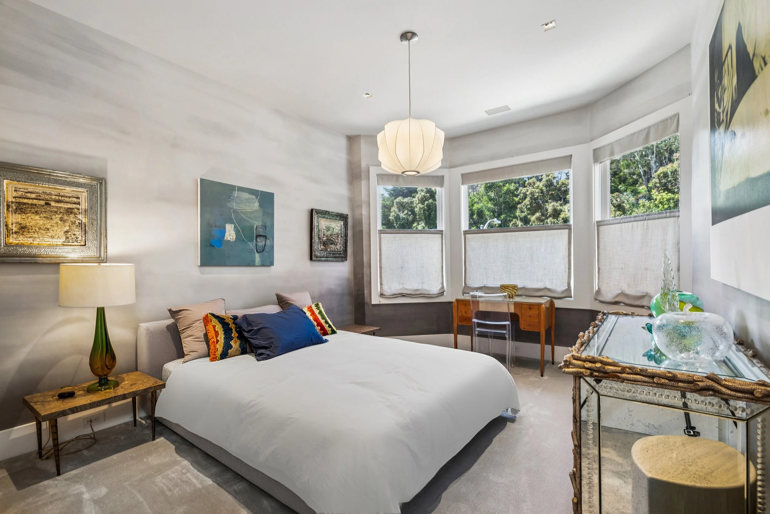 A bright, modern bedroom with a white bed, colorful pillows, a green and amber table lamp, wall art, large windows with roller shades, a wooden desk, and a glass-topped table with decorative glassware.