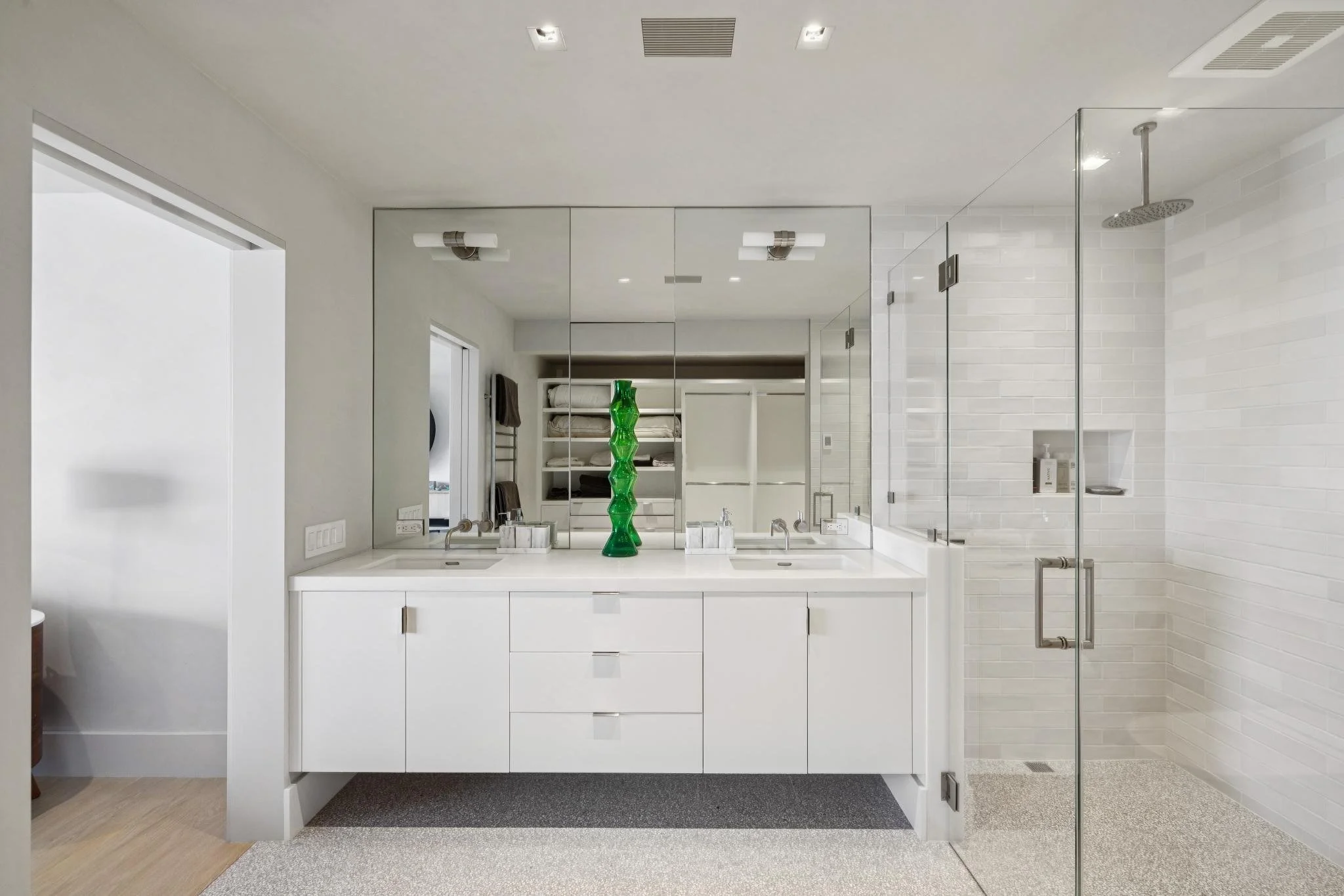 Modern bathroom with white vanity, large mirror, double sinks, green decorative vase, walk-in shower with glass door, white subway tiles, and gray and beige textured floor.