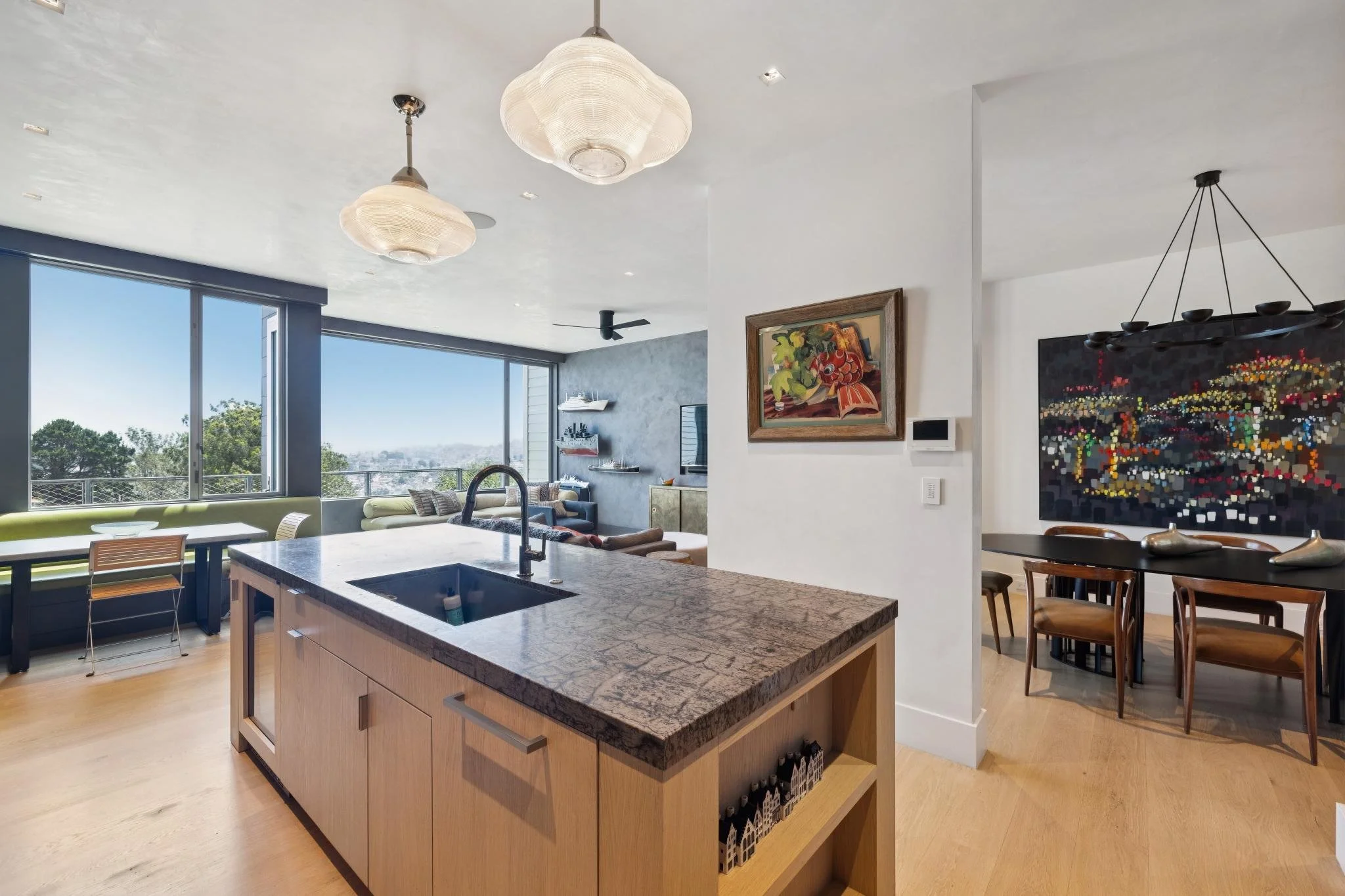 Open-concept modern living space with large windows, kitchen island with a sink, art on the walls, and a dining area.