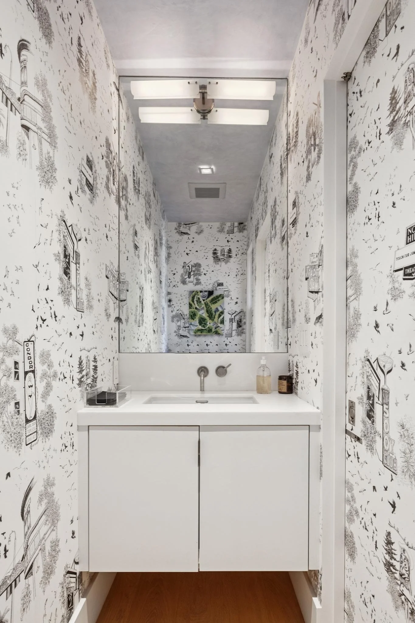 A small bathroom with wallpaper covered in black and white illustrations of trees, birds, signs, and landscape scenes. There's a white sink vanity with a modern tap, a hand soap dispenser, and decorative items on the counter. The ceiling has a contem