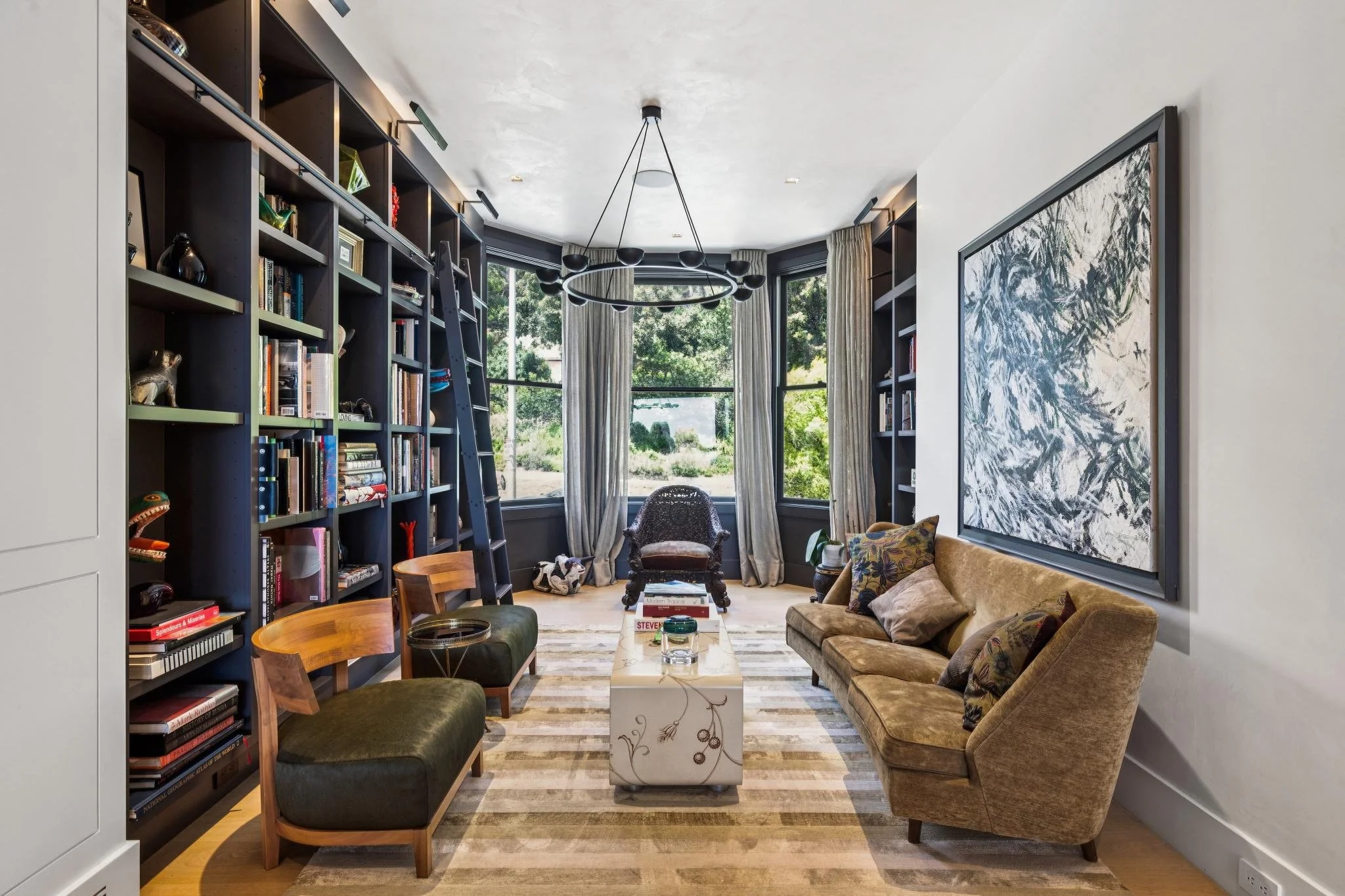 Living room with large black bookshelf, round chandelier, large windows with curtains, beige sofa, armchair, and coffee table.