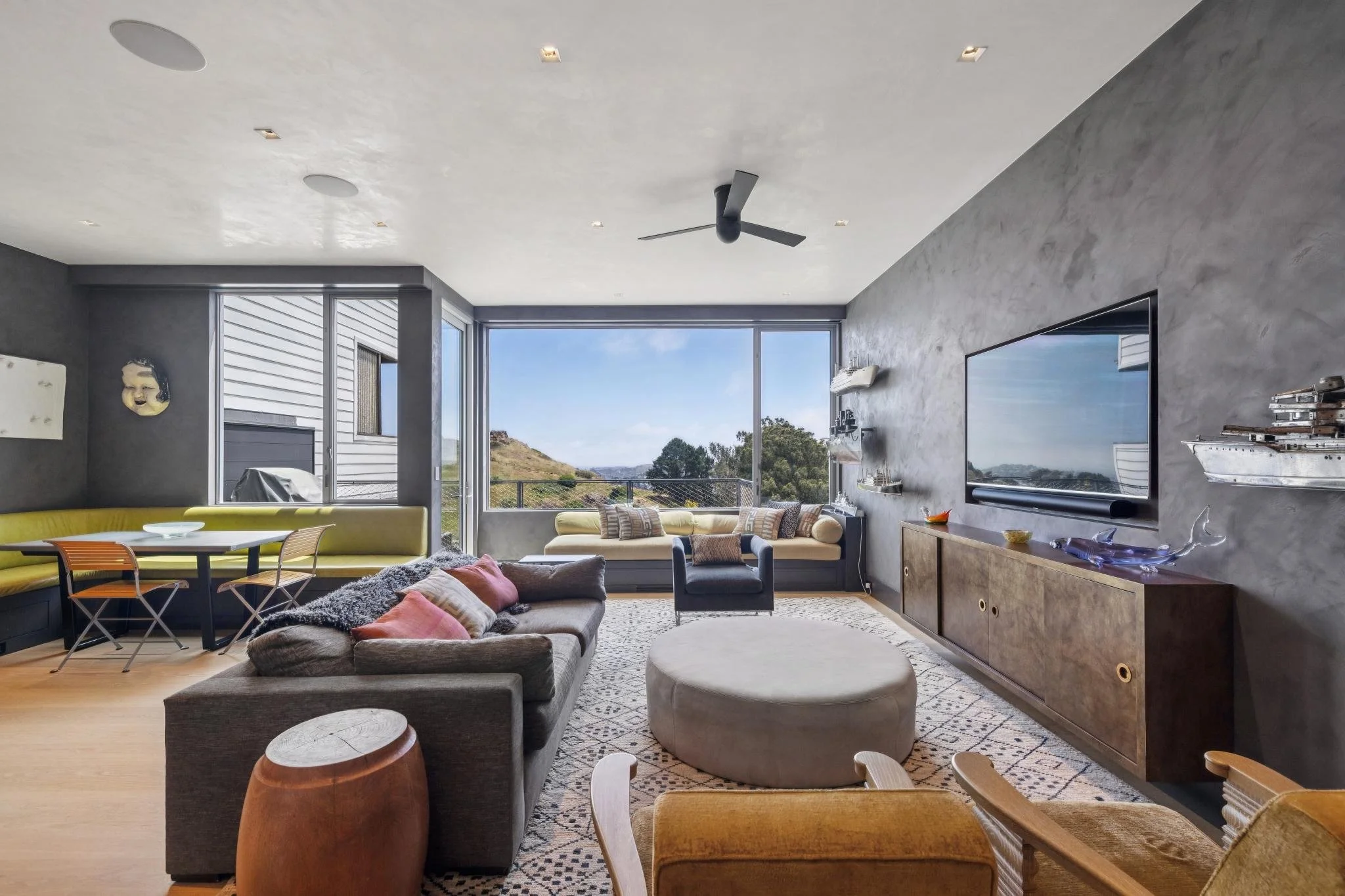 Modern living room with large window showing a view of hills and trees, featuring a gray sectional sofa, yellow banquette, a gray armchair, a large wall-mounted television, and various decorative items.