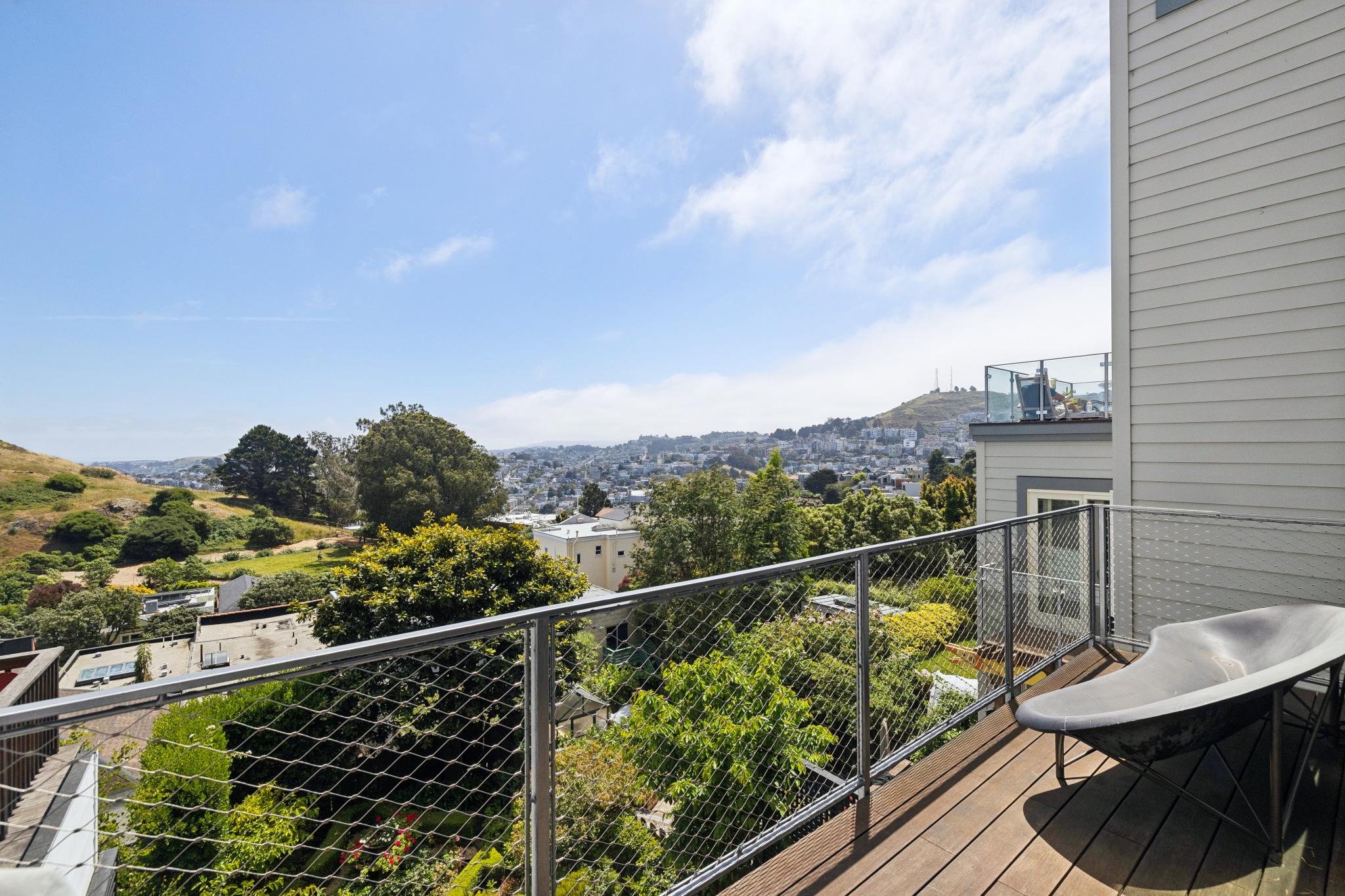 A balcony with wooden flooring and metal railing overlooking a green landscape with trees, houses, hills, and a partly cloudy sky.
