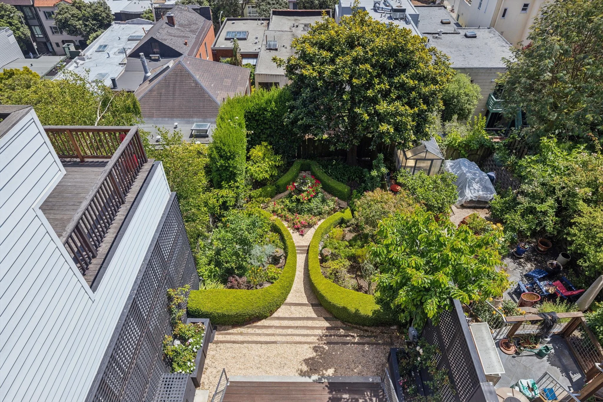 A bird's eye view of a backyard garden with a pathway, lush trees, flower beds, and surrounding houses with flat rooftops.