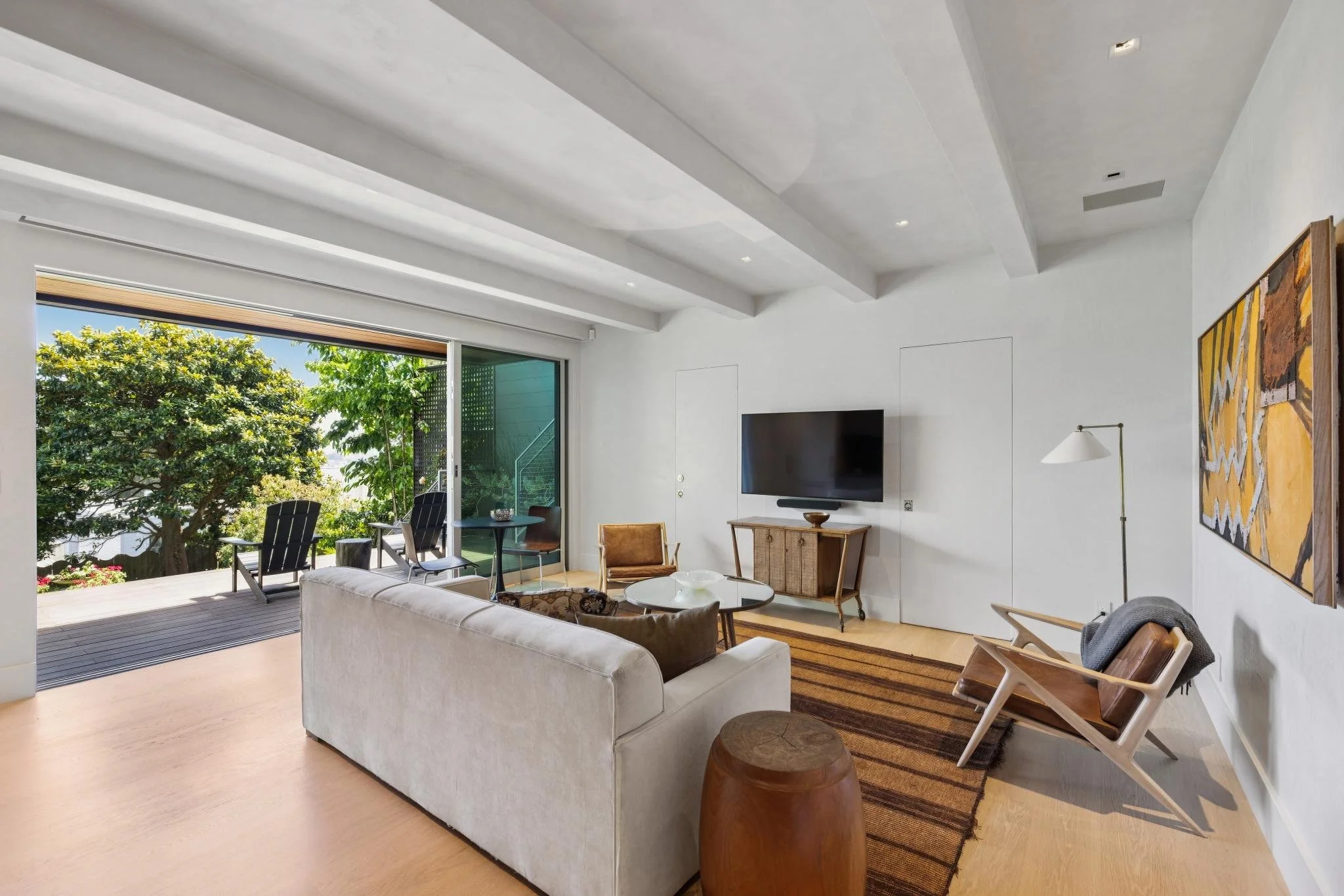 A living room with a white sofa facing a wall-mounted flat-screen TV, a wooden cabinet beneath it, and a colorful painting on the wall. There is an armchair with a blanket draped over it, a small round table, and a wooden stool. The room opens to a d