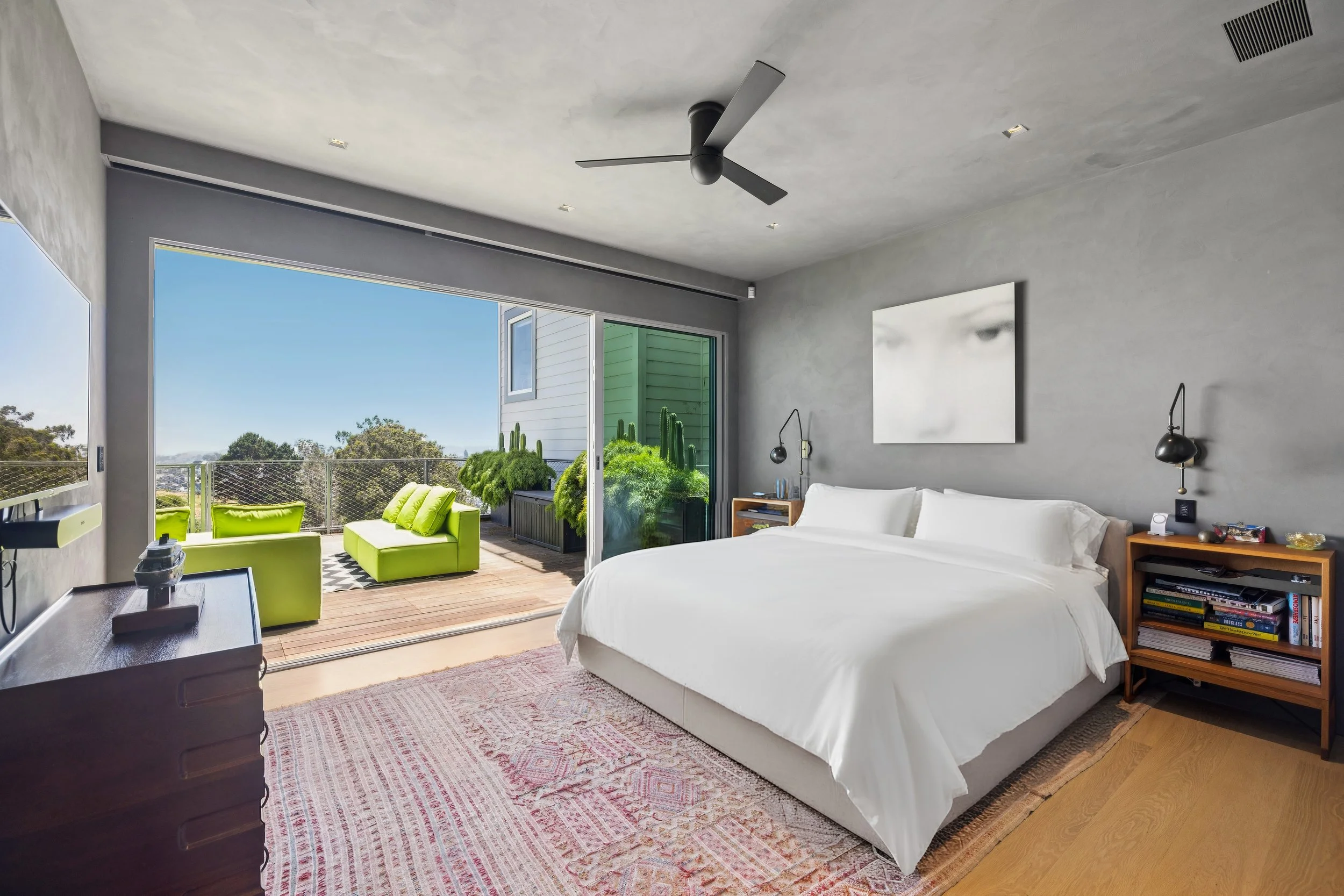 Modern bedroom with large sliding glass door opening to a balcony with green outdoor seating and plants, white bed with bedside tables, a painting on the wall, black wall-mounted lights, a ceiling fan, and a patterned rug.