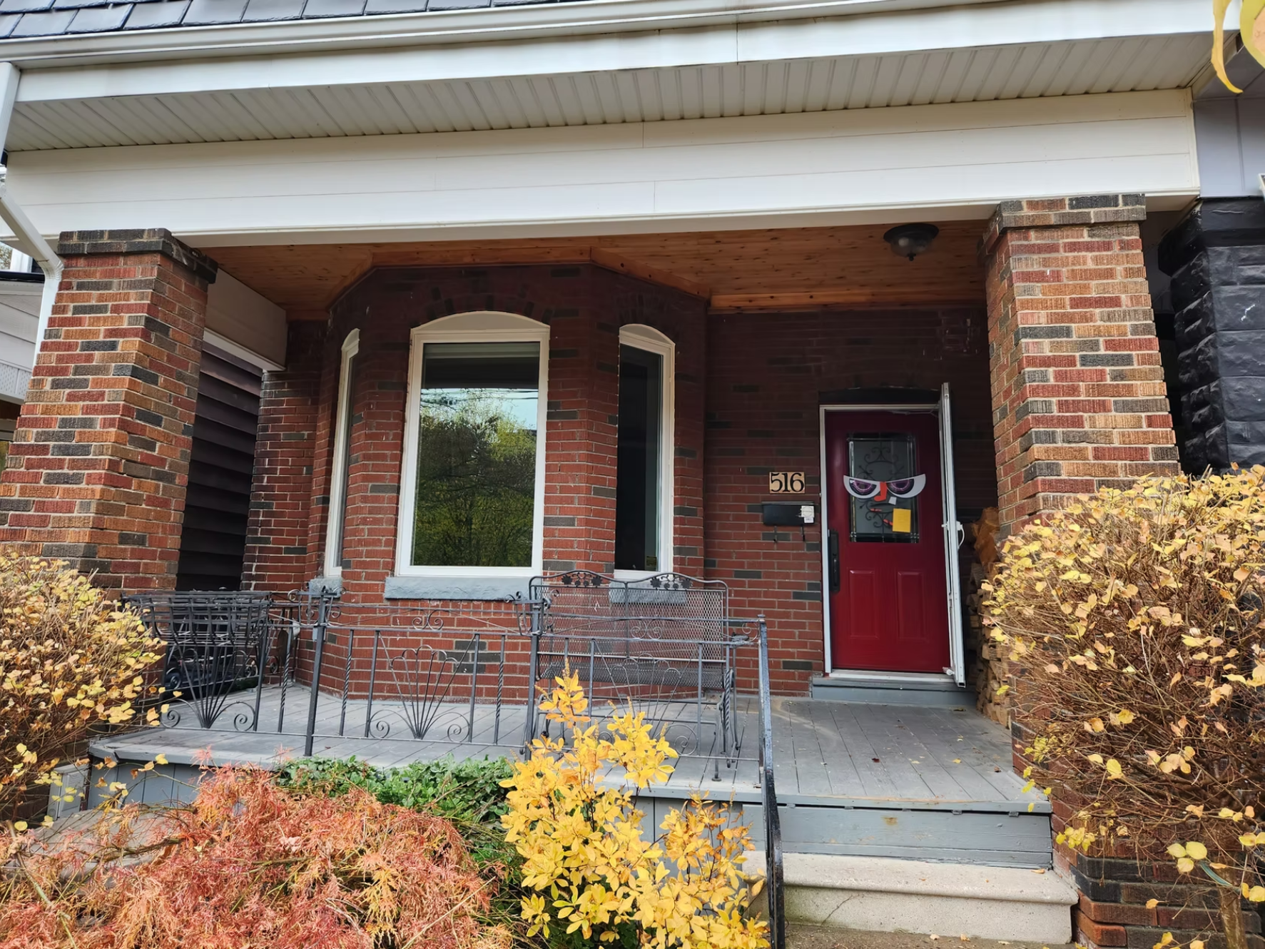 Front porch of a brick house with a red door decorated with Halloween-themed paper glasses. The porch has black metal railings, and there are bushes with yellow and red leaves in the front yard. The house number, 516, is visible on the wall next to the door.