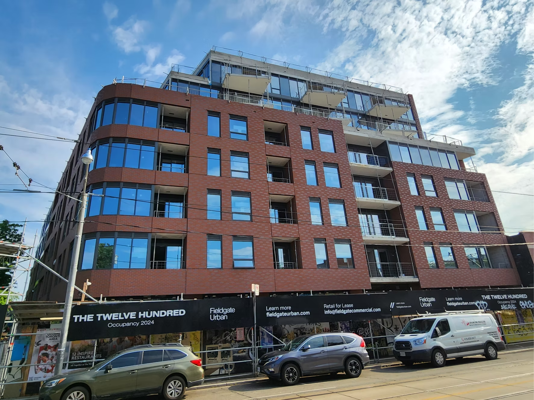 Modern multi-story building under construction with brick and glass facade, construction scaffolding at the top, cars parked in front, and a black banner that reads 'The Twelve Hundred'.