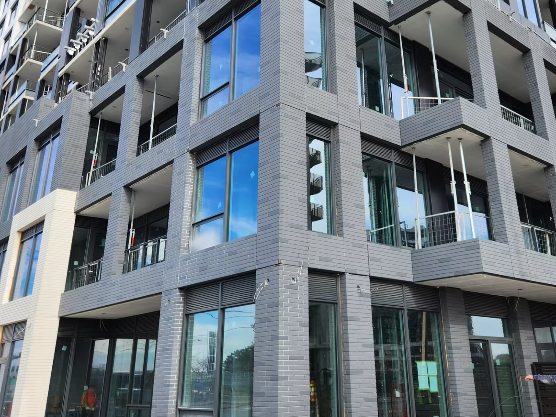A modern multi-story building with gray brick exterior, large windows reflecting the sky, and balconies with metal railings. Construction is ongoing, with some scaffolding and building fixtures visible.