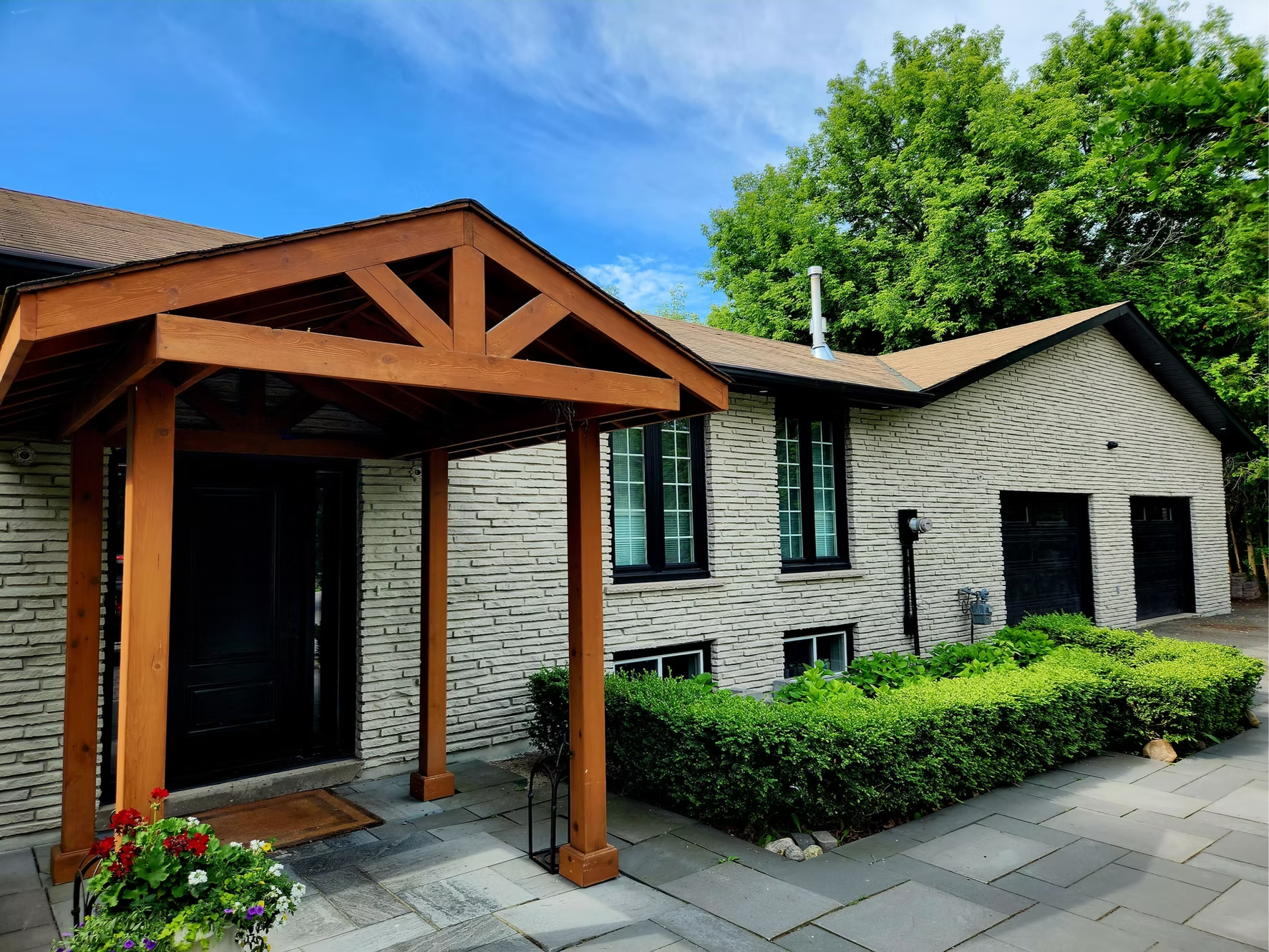 Exterior view of a residential house with a brick facade, green bushes, a paved walkway, and a wooden porch structure with a lattice design, under a partly cloudy sky.
