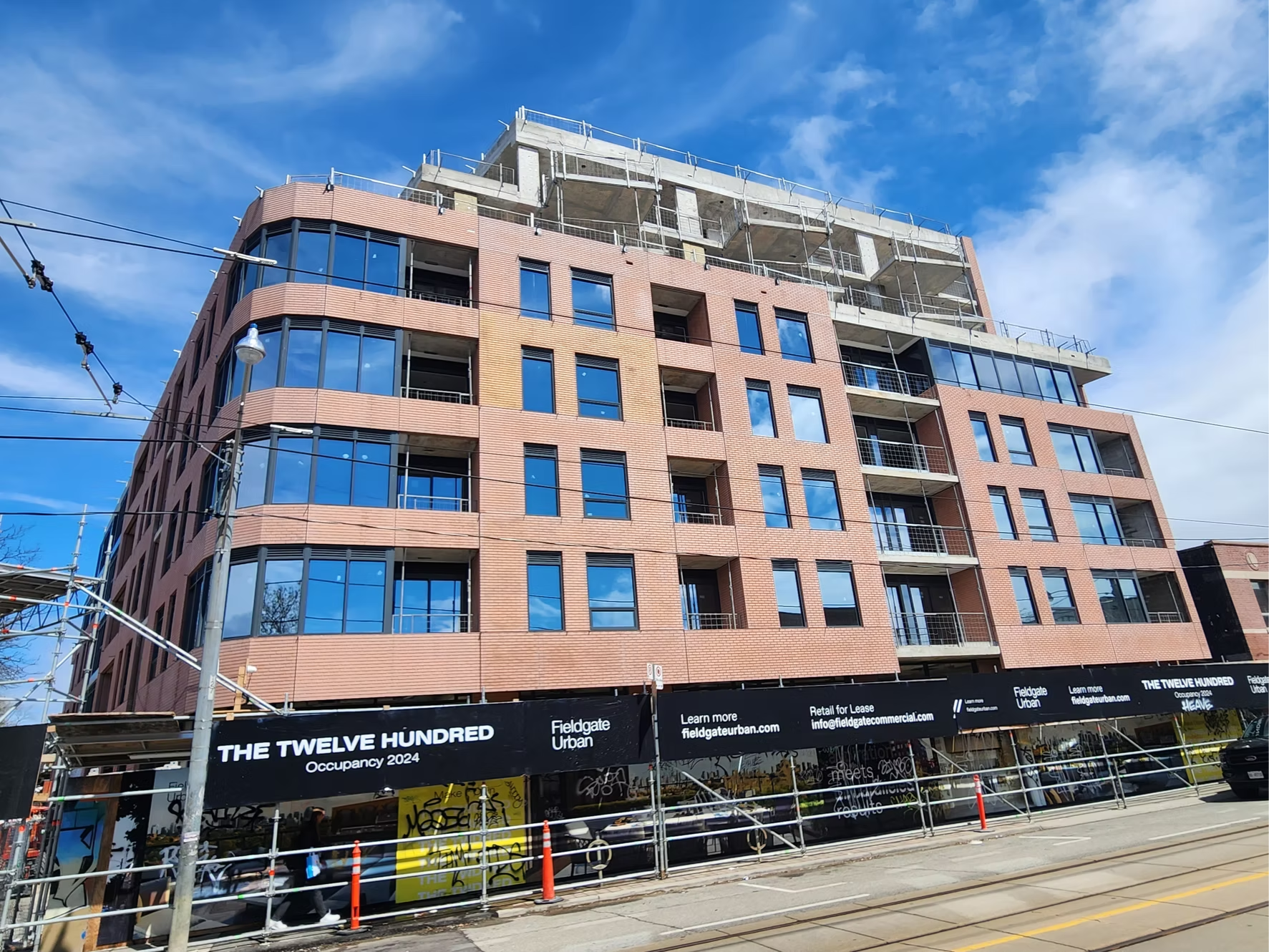 A multi-story building under construction with a brick facade and large blue-tinted windows. There is construction scaffolding and equipment around the building, with a banner indicating it is called 'The Twelve Hundred' for occupancy in 2024, located in an urban area with tram tracks and overhead wires.