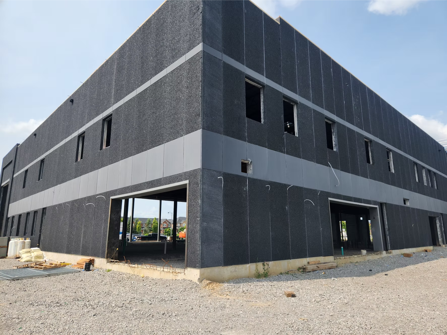 A multi-story building under construction with black and gray exterior walls, open window spaces, and construction materials on the ground, set against a partly cloudy sky.