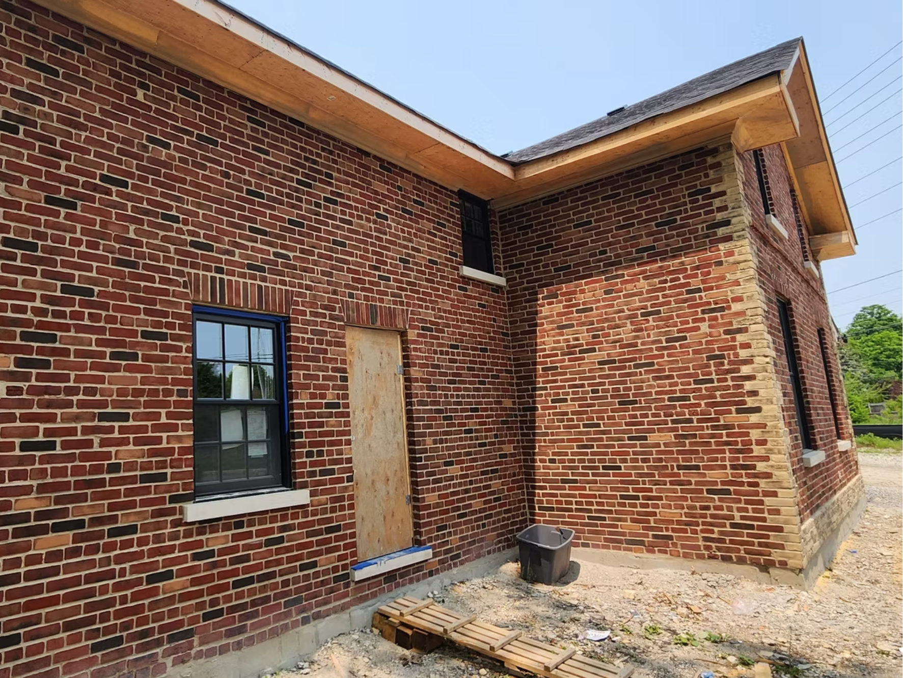 A brick house under construction with windows and wooden roof overhang, construction materials, and a trash bin outside.