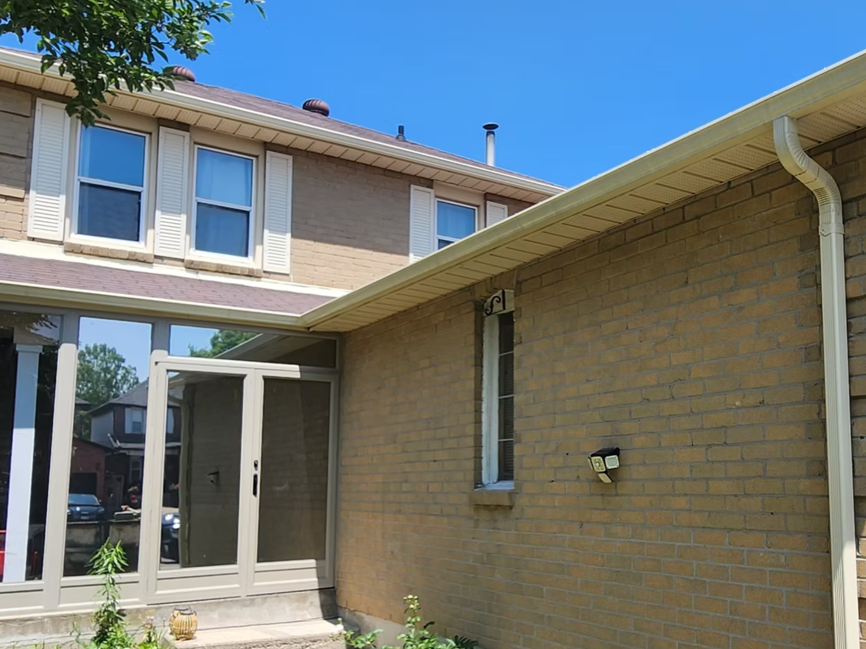 The image shows the exterior of a two-story brick house with white window shutters, a beige roof with vents, and a chimney with a pipe. There is a screened porch with white framing and a small garden area with plants in the foreground. The sky is clear and blue.