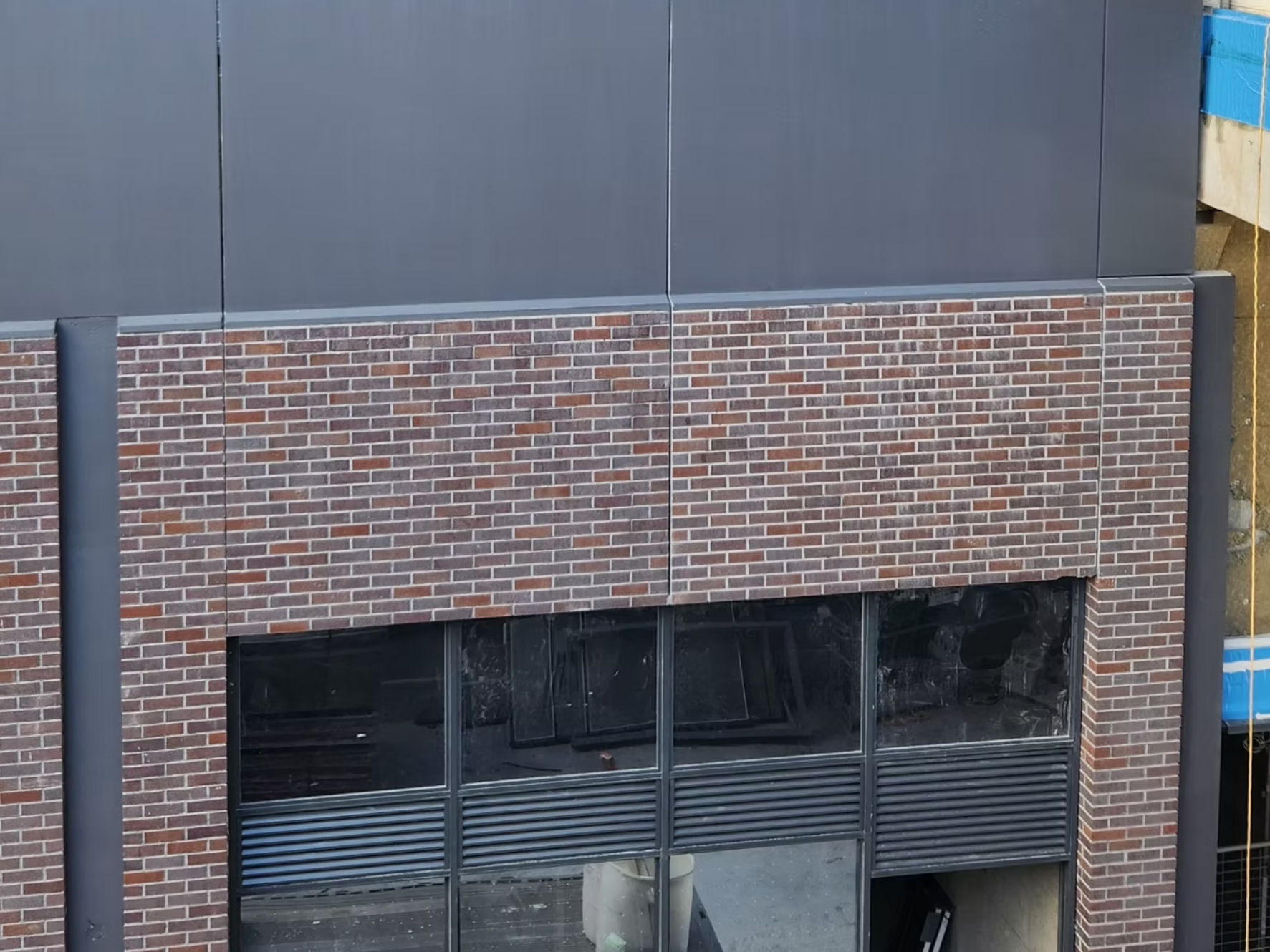 Close-up of a building's exterior wall with a combination of brick and gray metal panels, featuring large glass windows and vertical metal poles on the sides.