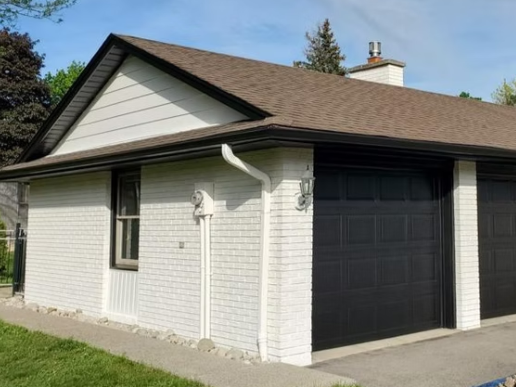 Front view of a house featuring a white brick exterior, black garage door, brown shingle roof, and a white gutter system, with a window on the side and a small patch of green grass in the front.