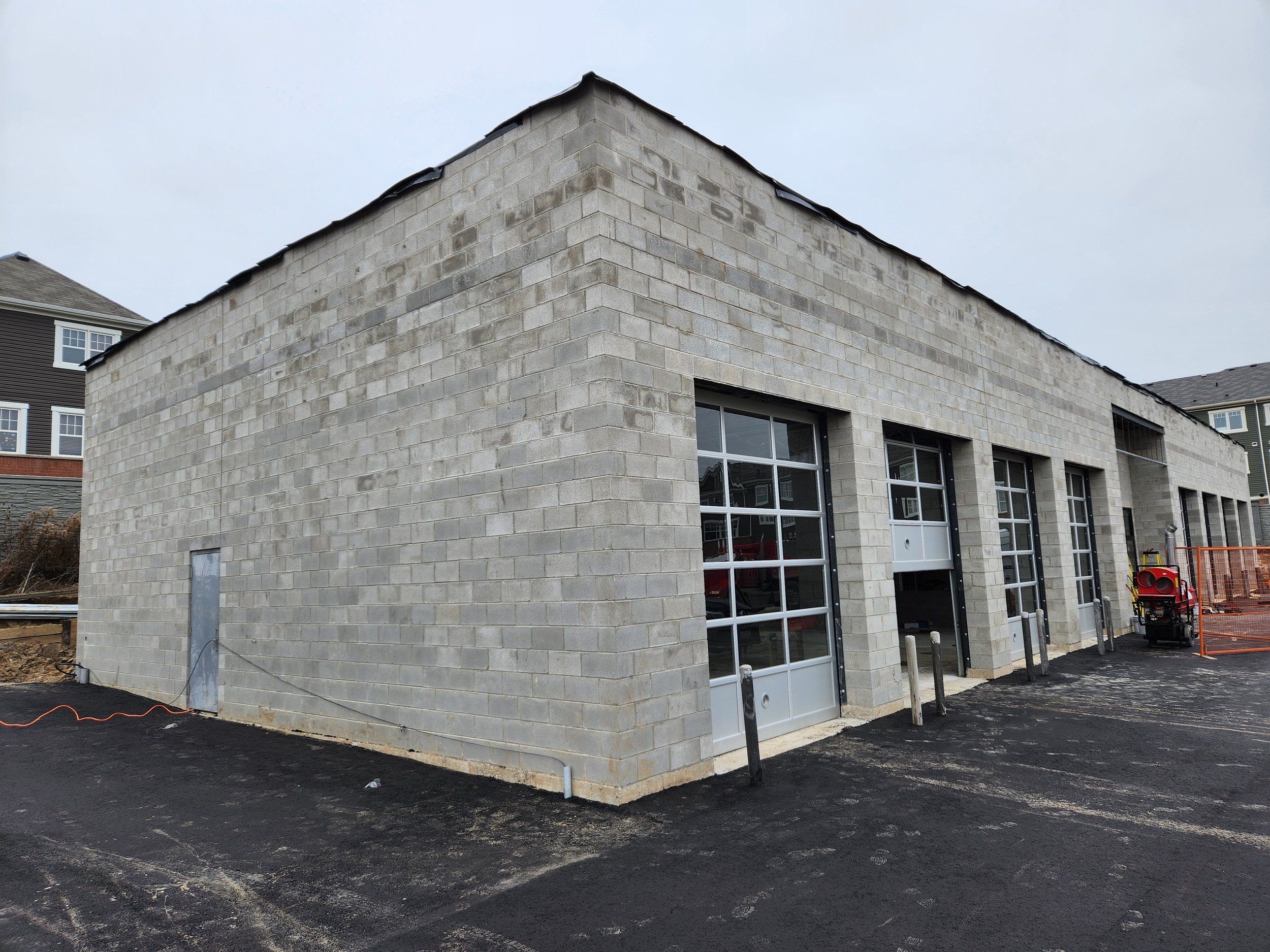 A modern building under construction with a cinder block exterior and large glass windows. There are construction materials and equipment around the building, and the ground is freshly paved.