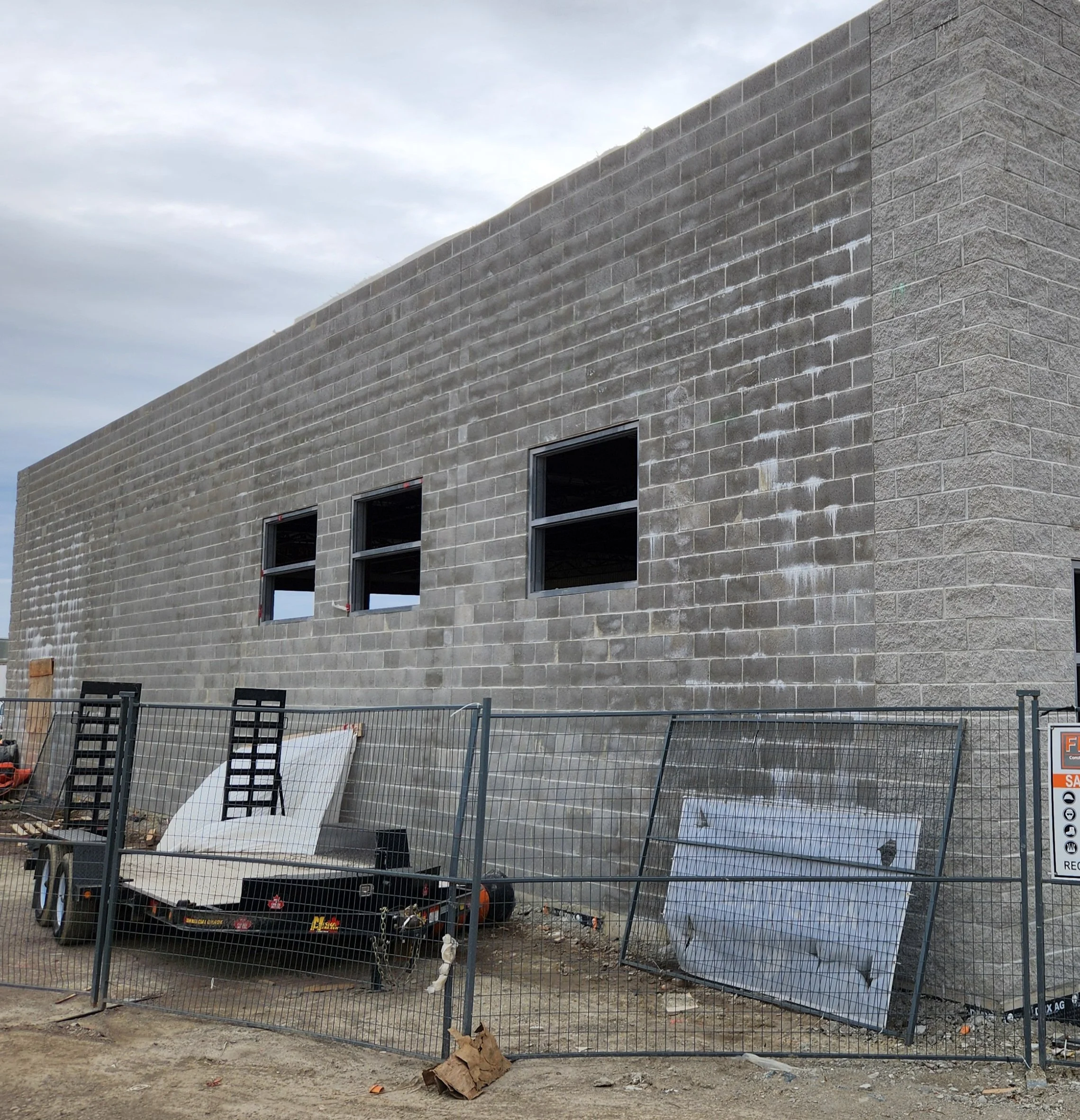 Construction site with a gray brick building and open windows, surrounded by a metal fence, with construction materials and temporary signs on the ground.