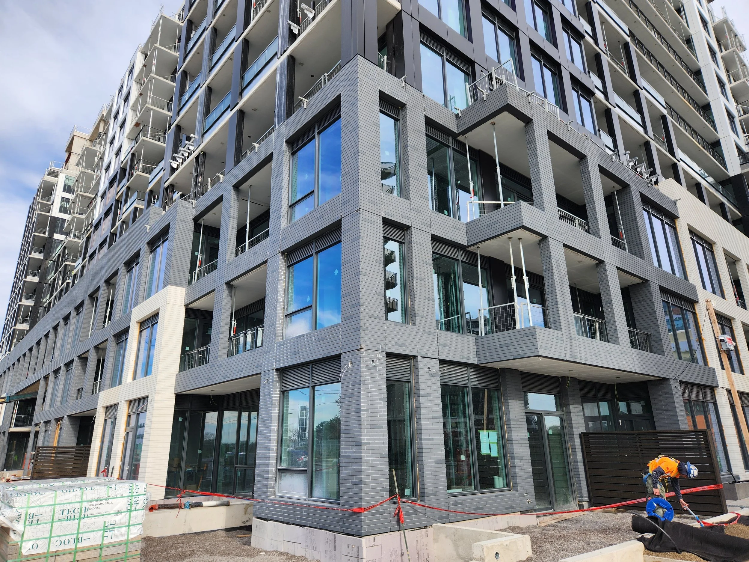 Modern multi-story building under construction with glass windows and balconies, construction workers working at the base, and construction materials visible in the foreground.