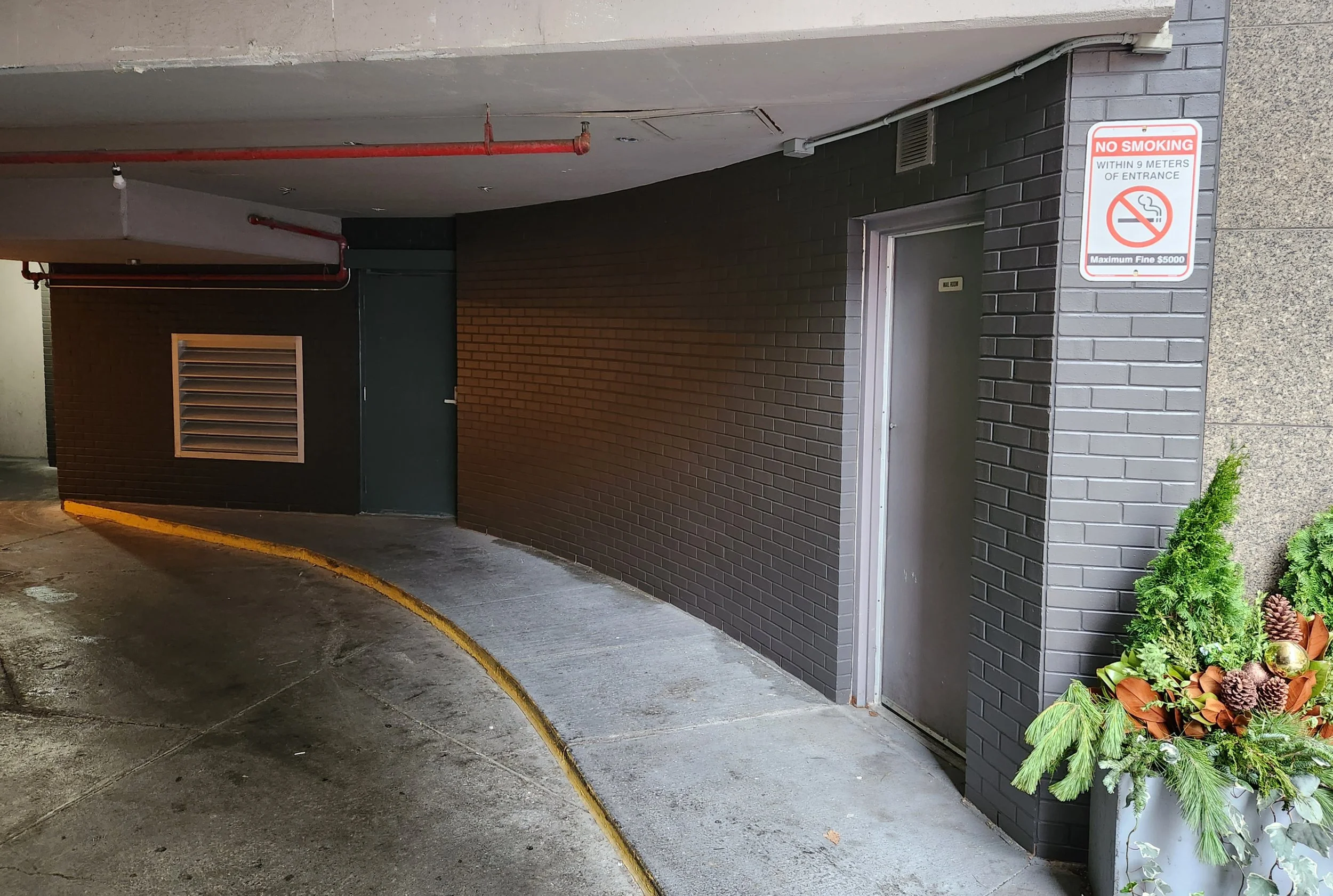 A dark gray brick wall with a gray door, a vent, a sign that says 'No Smoking,' red pipes on the ceiling, and a small display of green and brown plants at the corner.