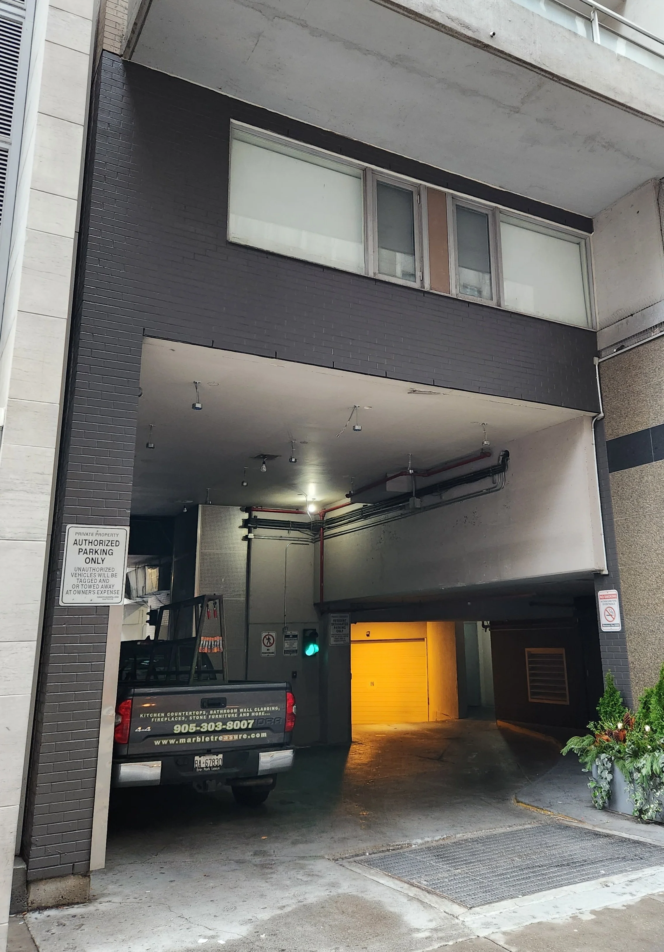 Underground parking garage entrance with a black truck parked, yellow garage door open, and signage indicating authorized parking only. Pipes and lighting on the ceiling, with some decorative plants on the right.