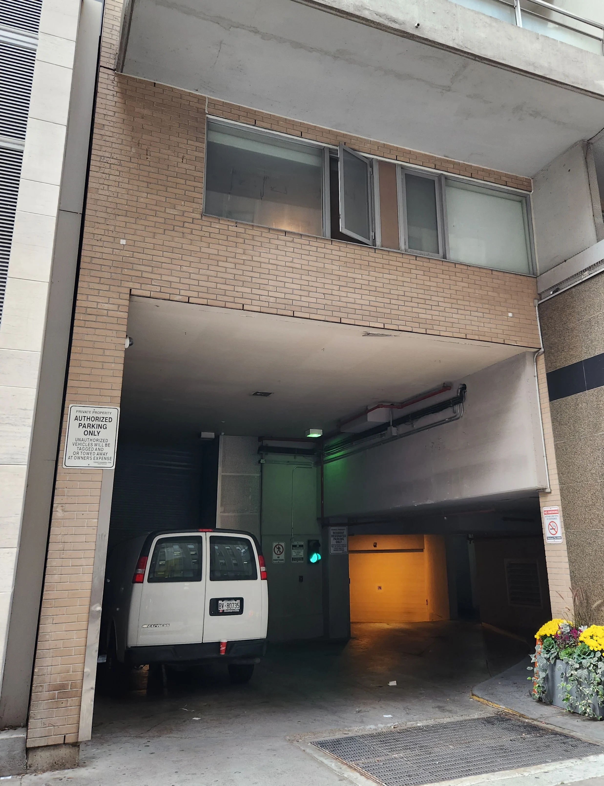 A parking garage entrance with a white van parked outside. The garage has brick walls and a roll-up door, with signs indicating authorized parking only. There are some flowers in a planter on the right side.
