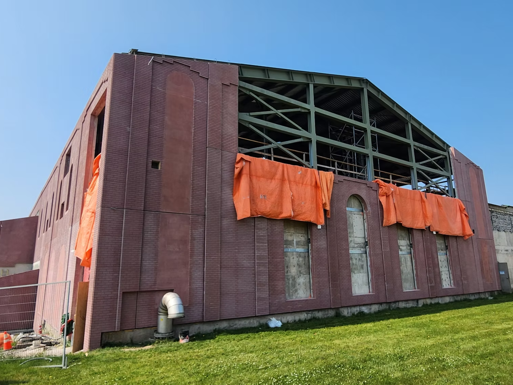 Building under construction with red brick walls, scaffolding, and orange tarps hanging on the exterior, against a clear blue sky and green grass.