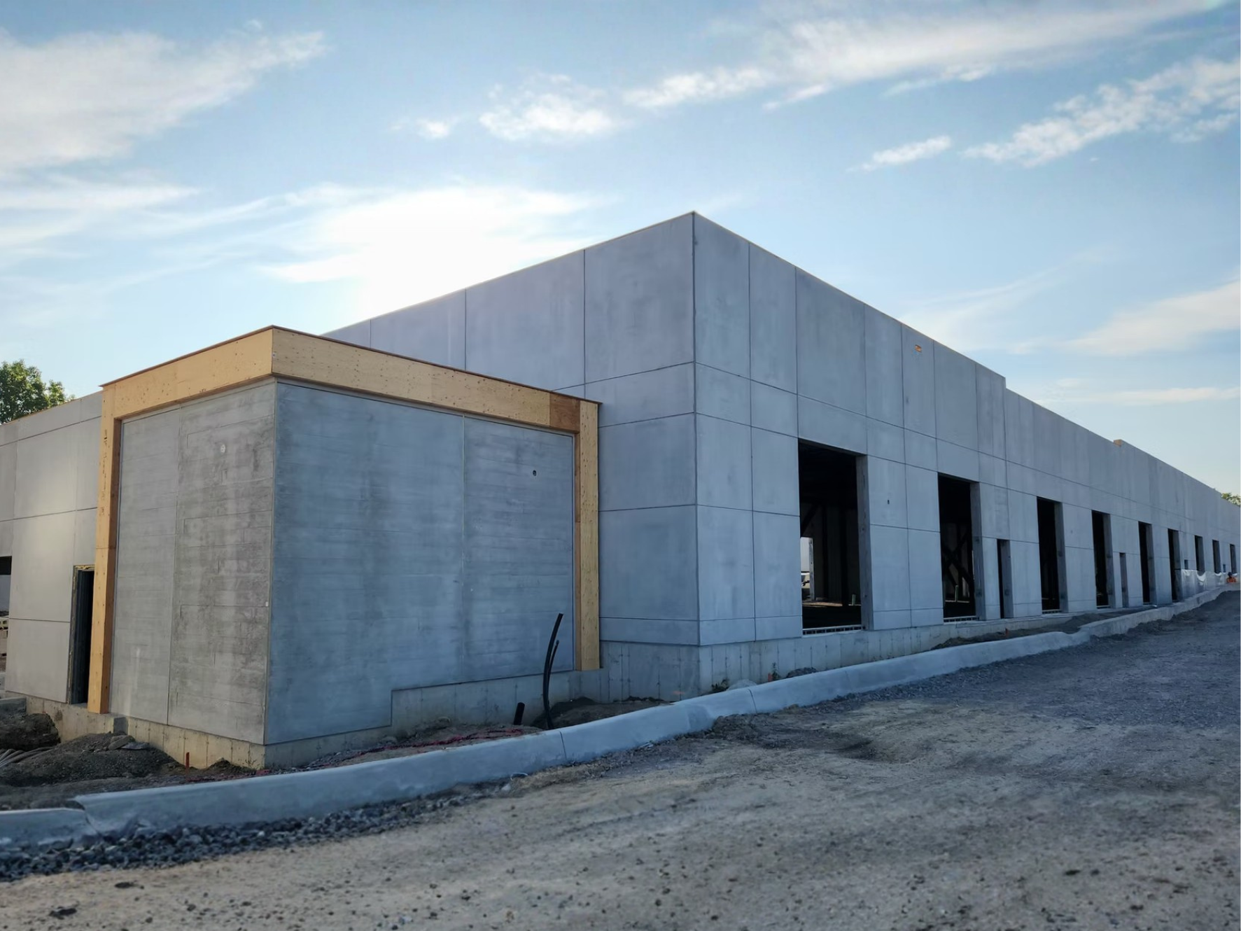 A building under construction with concrete walls and multiple large rectangular openings for windows or doors. The building has a partial wooden frame around one section. The sky is clear with some clouds, and there is construction material on the ground in front of the building.