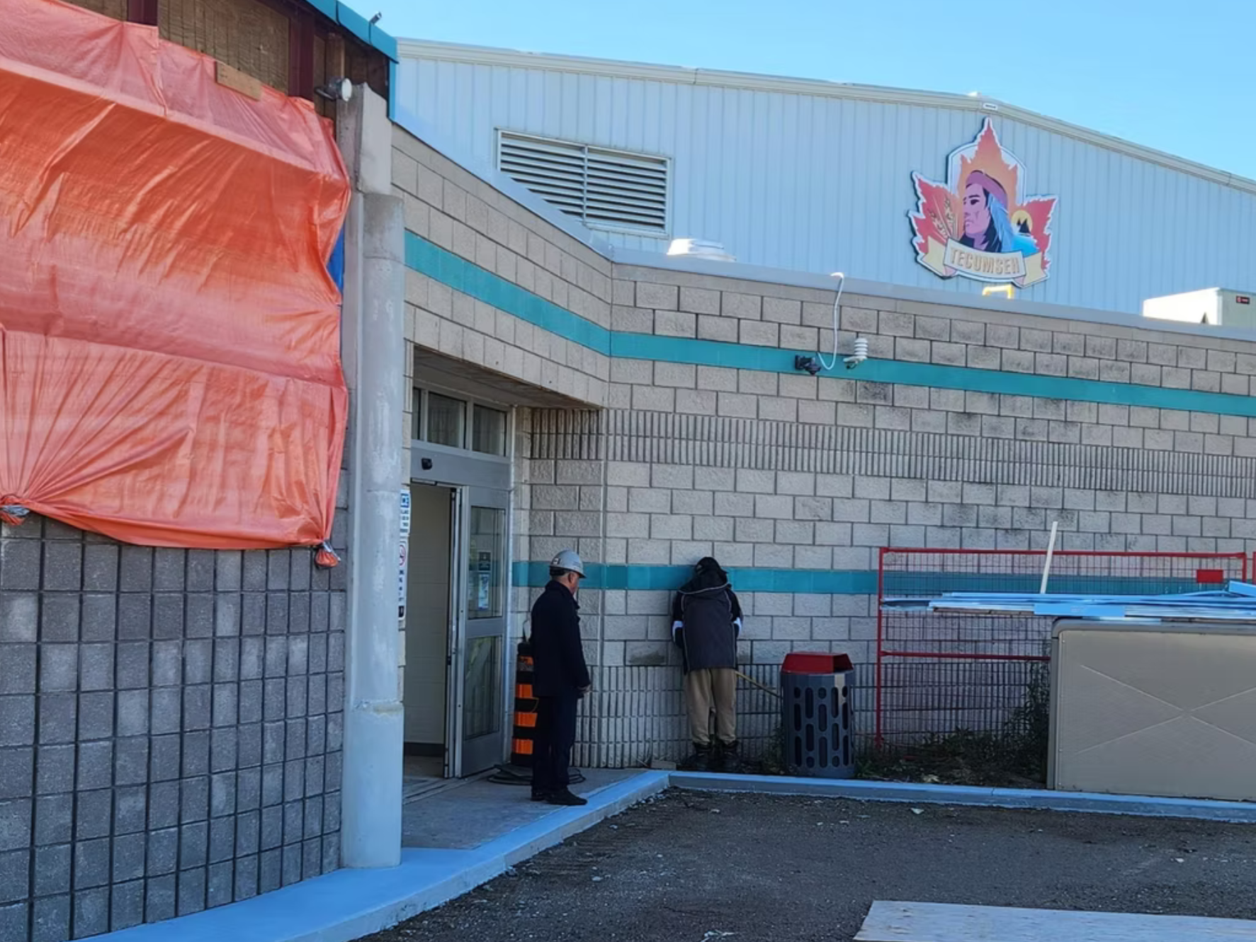 Construction workers standing near the entrance of a building under construction, with a mural of Tecumseh on the upper wall, and orange construction tarp covering part of the structure.