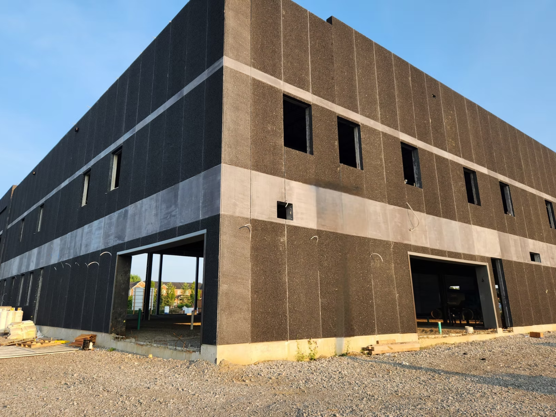 Under construction two-story building with black exterior walls, multiple window openings, and a large open garage or entrance on the ground floor. The building is surrounded by gravel and construction materials, with a clear blue sky above.