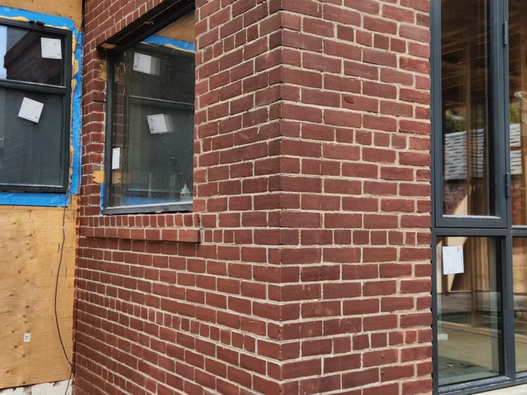 Close-up of a brick wall with a corner, showing a window with a black frame and partially installed blue tarp and wooden paneling on a building under construction.