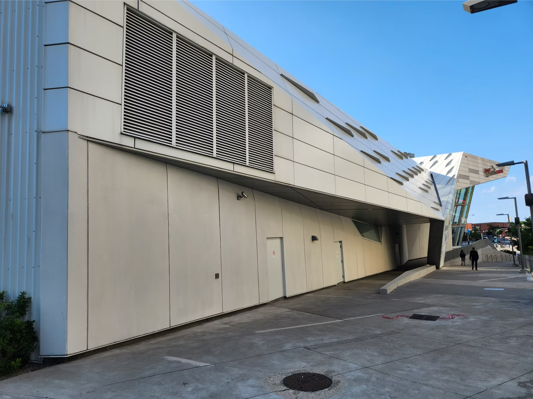 Modern building with sleek white exterior, large ventilation grille, angled architectural design, and glass windows. Sidewalk in front with two people walking and street lamps visible.