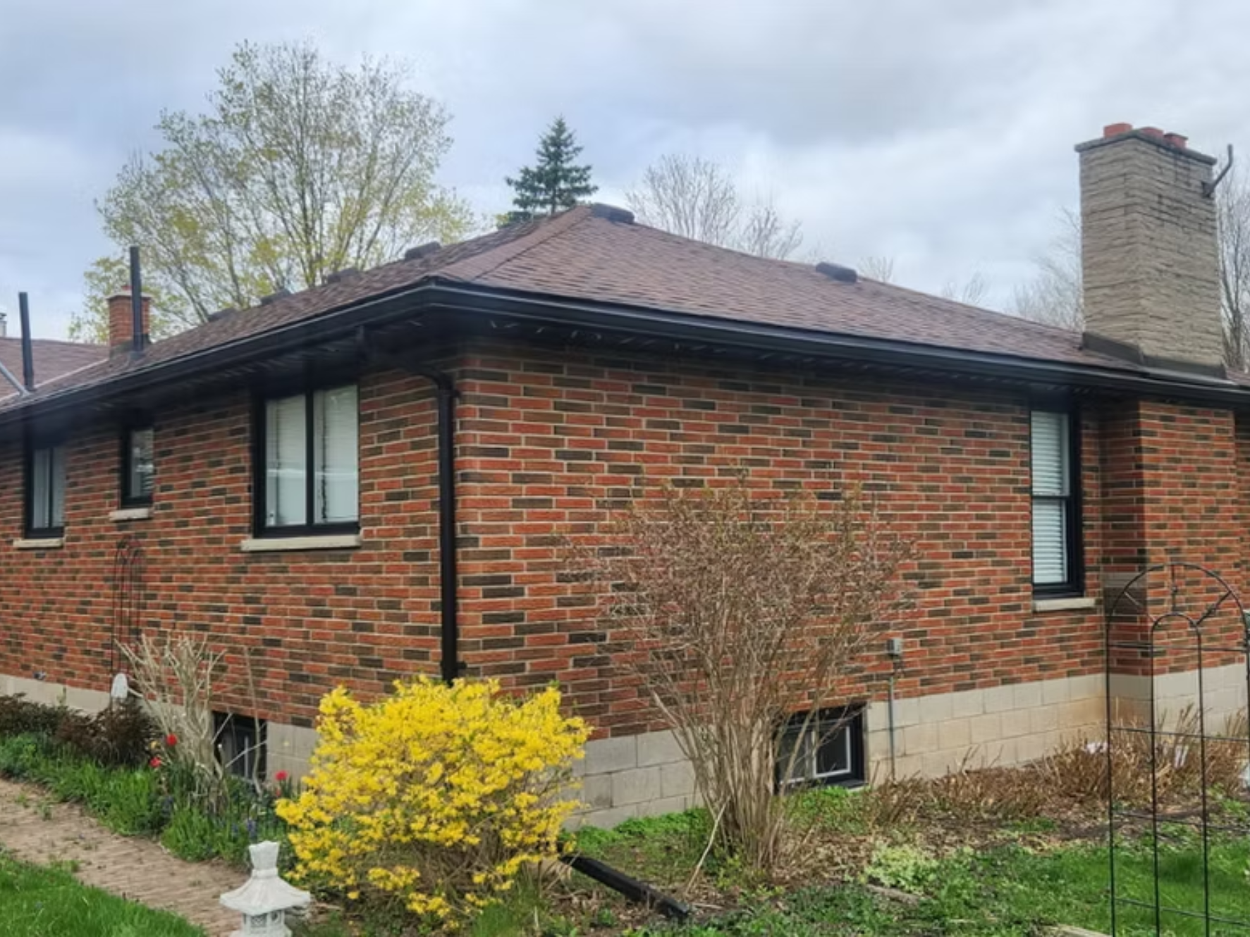 A two-story brick house with a sloped shingle roof, multiple windows with black frames, and a tall brick chimney. There is a garden with small trees, bushes, and yellow flowers in front of the house.